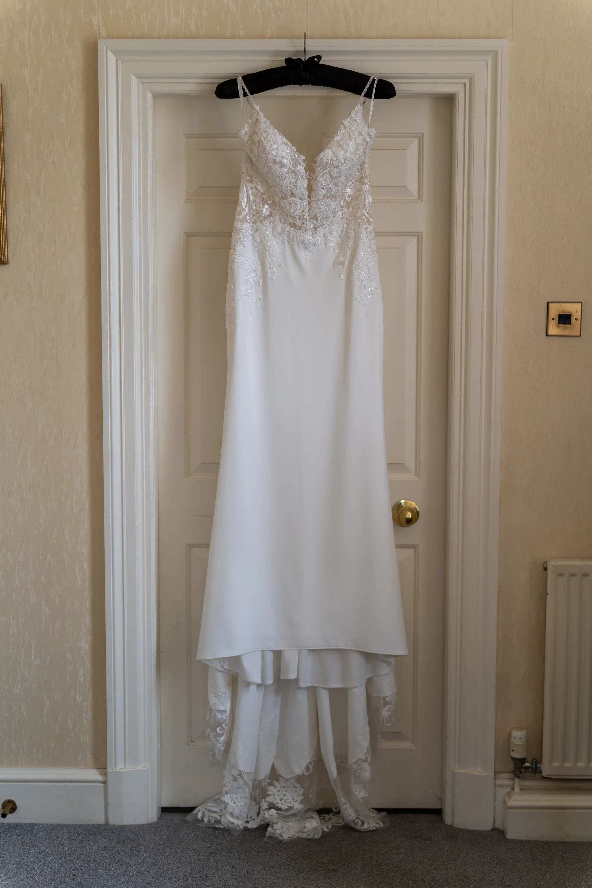 An intricate lace and crepe wedding dress hangs elegantly from a black hanger against a white panelled door during bridal preparations.