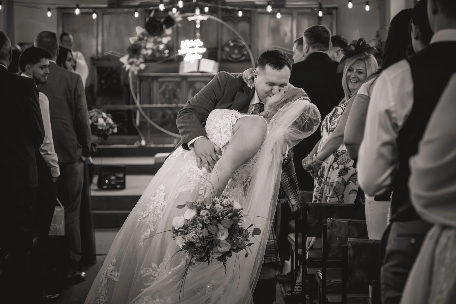 The newly married groom dips the bride in a passionate kiss at the altar of a church while guests watch from the pews.