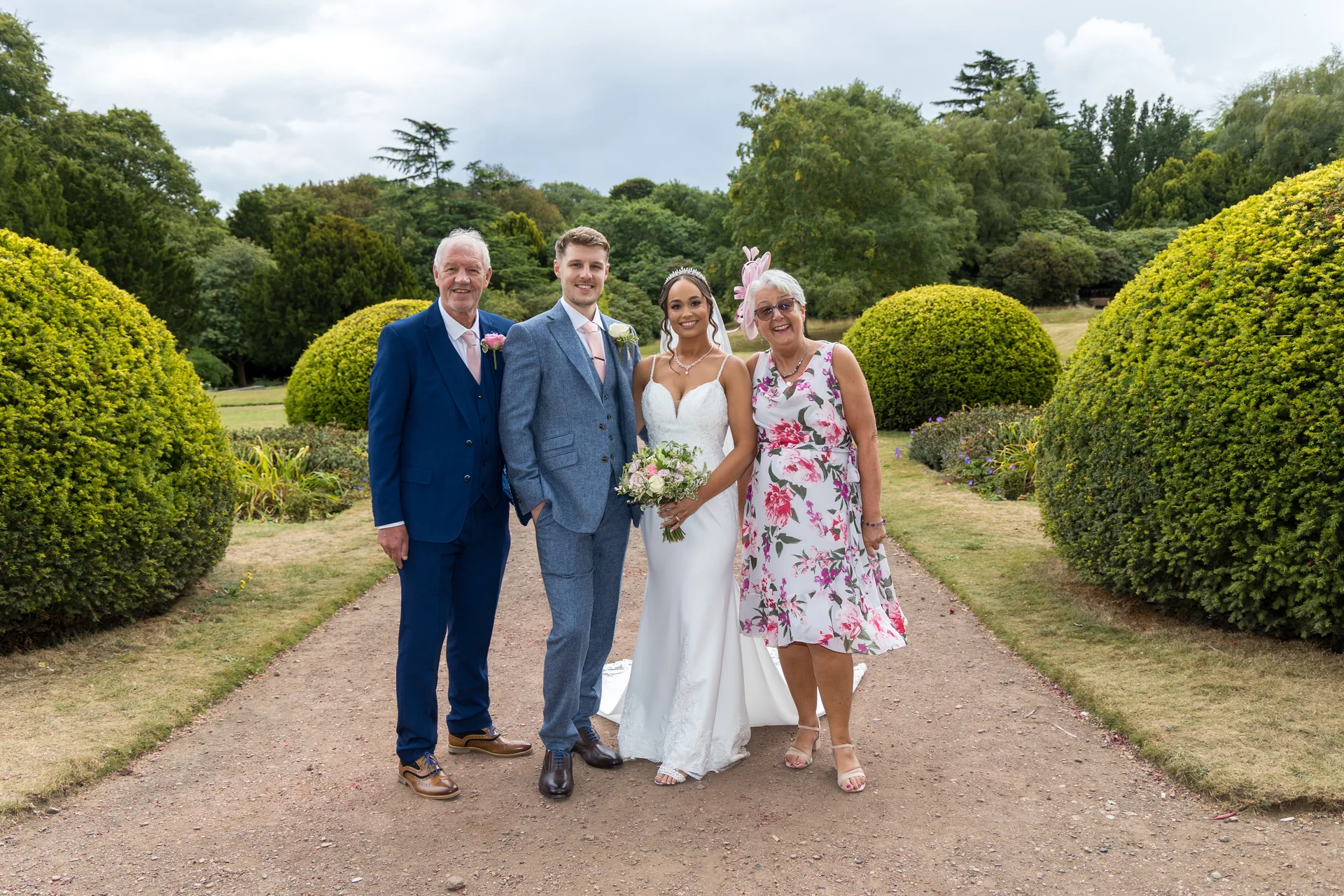 A newlywed couple poses outdoors on a garden path alongside their parents for a traditional formal wedding portrait framed by manicured topiary bushes.
