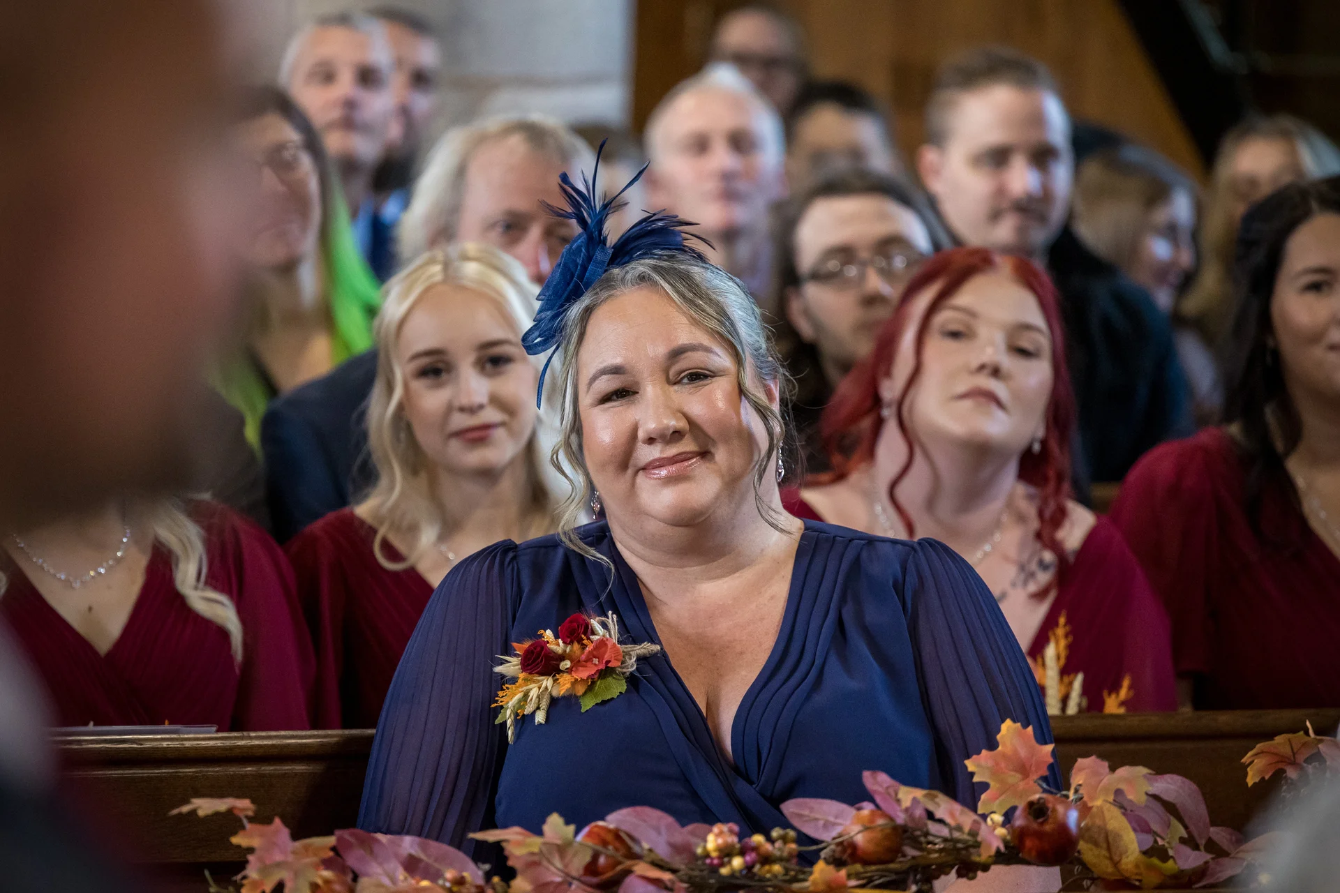 A smiling guest wearing a blue dress and fascinator sits among seated wedding attendees inside a church during a ceremony.