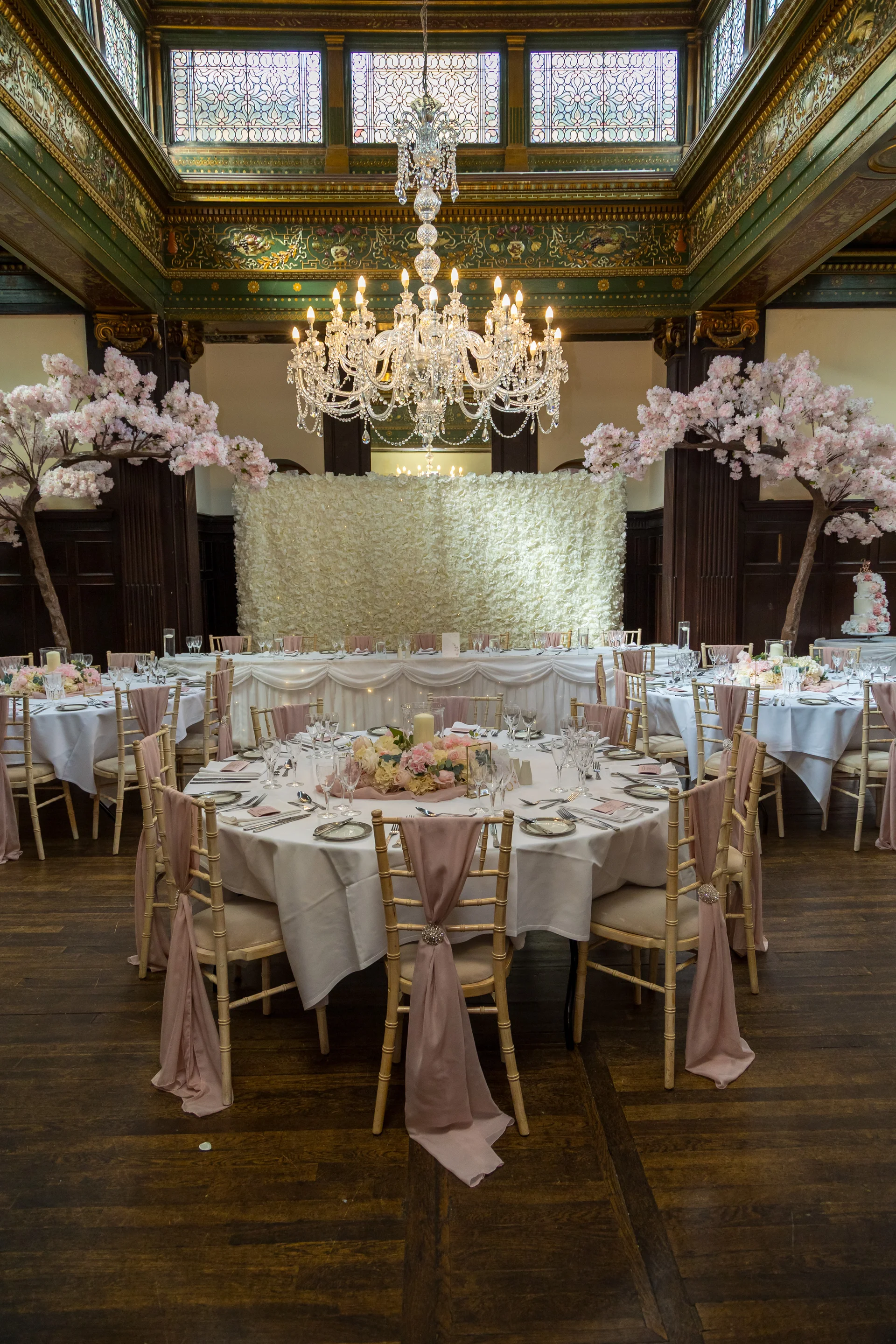 An elegantly decorated wedding reception ballroom features round tables set with white linens, dusty pink chair sashes, and floral centrepieces beneath a grand crystal chandelier and stained glass windows.