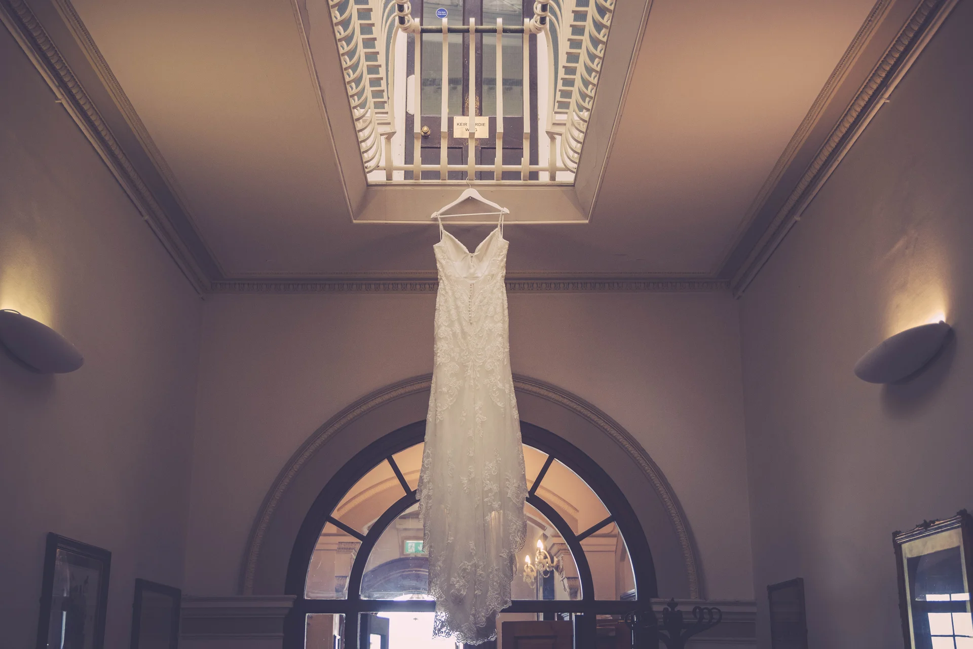 An intricate lace wedding dress hangs elegantly from a railing within an indoor venue featuring grand architectural arches and warm lighting.