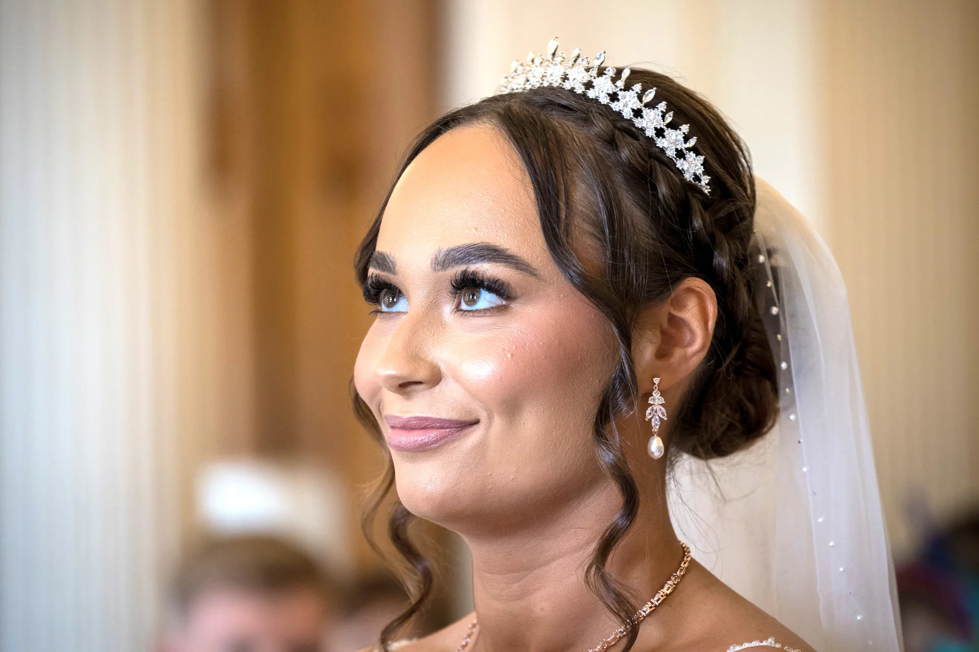 Close up portrait of a smiling bride wearing a sparkling tiara, drop earrings, and an elegant updo with face-framing tendrils.