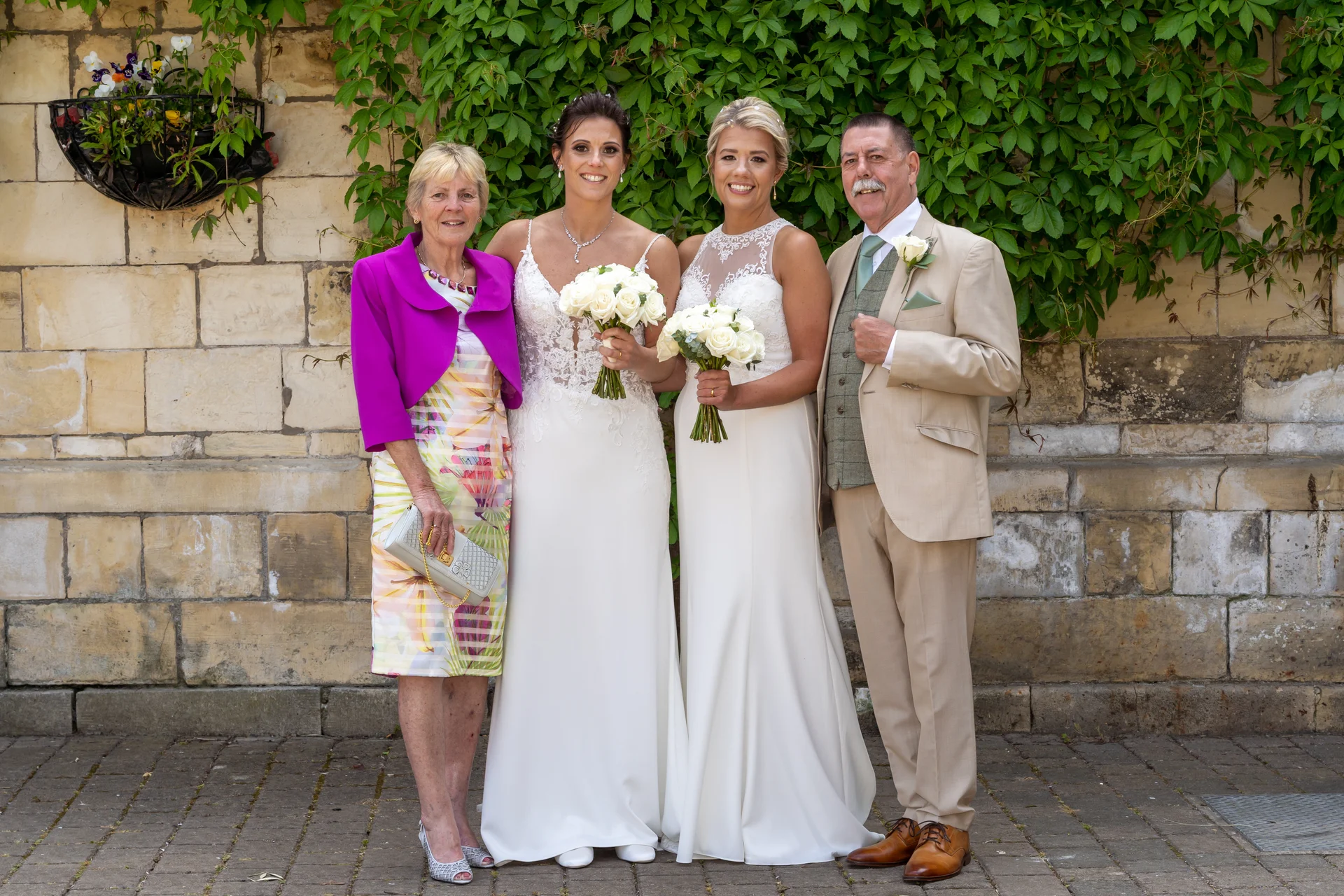 Two brides in white wedding dresses pose alongside a smiling older woman and a man in a beige suit against a rustic stone wall covered in green ivy.