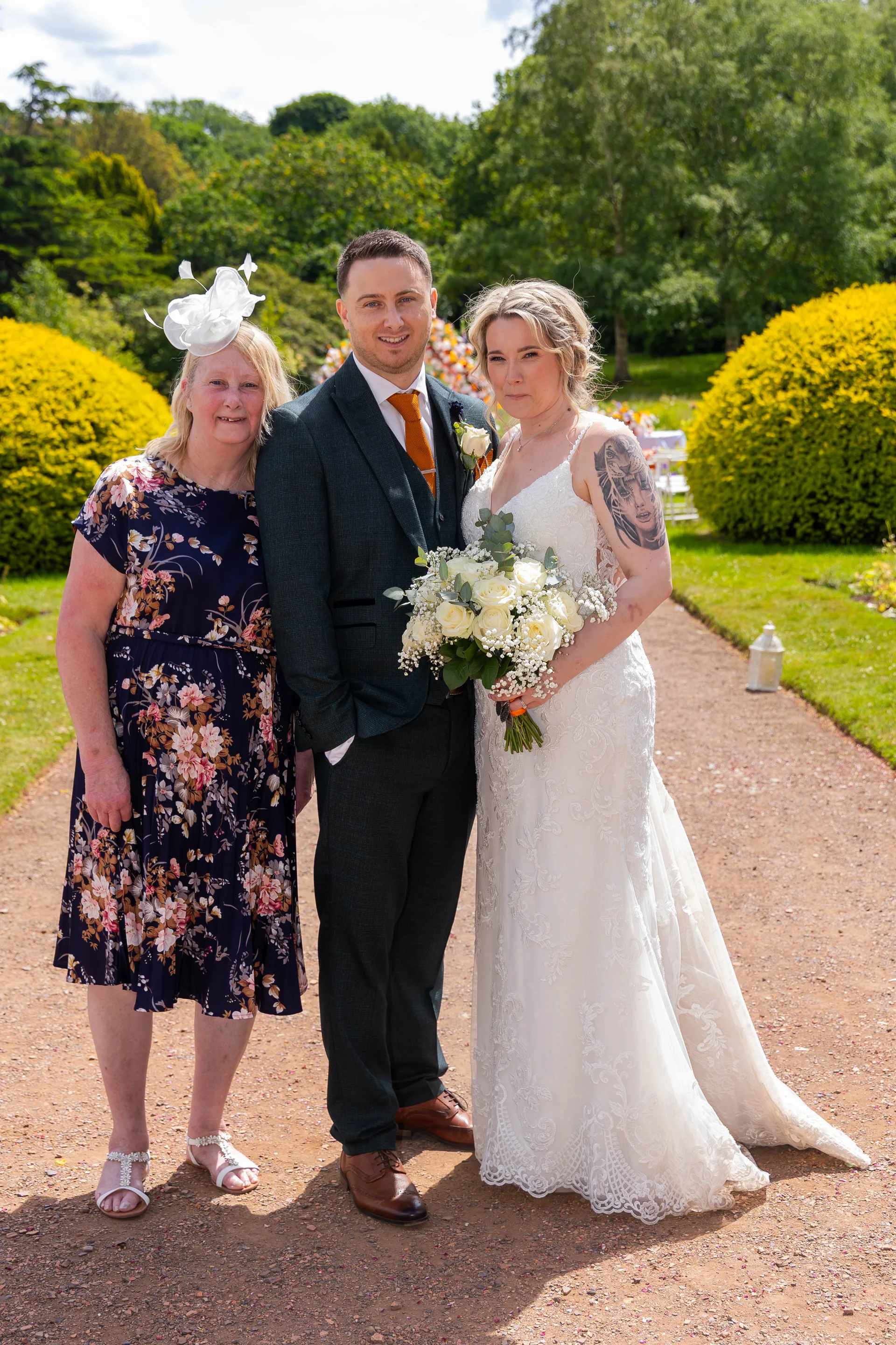 A newlywed couple poses outdoors in a sunlit garden alongside a guest wearing a floral dress and white fascinator.