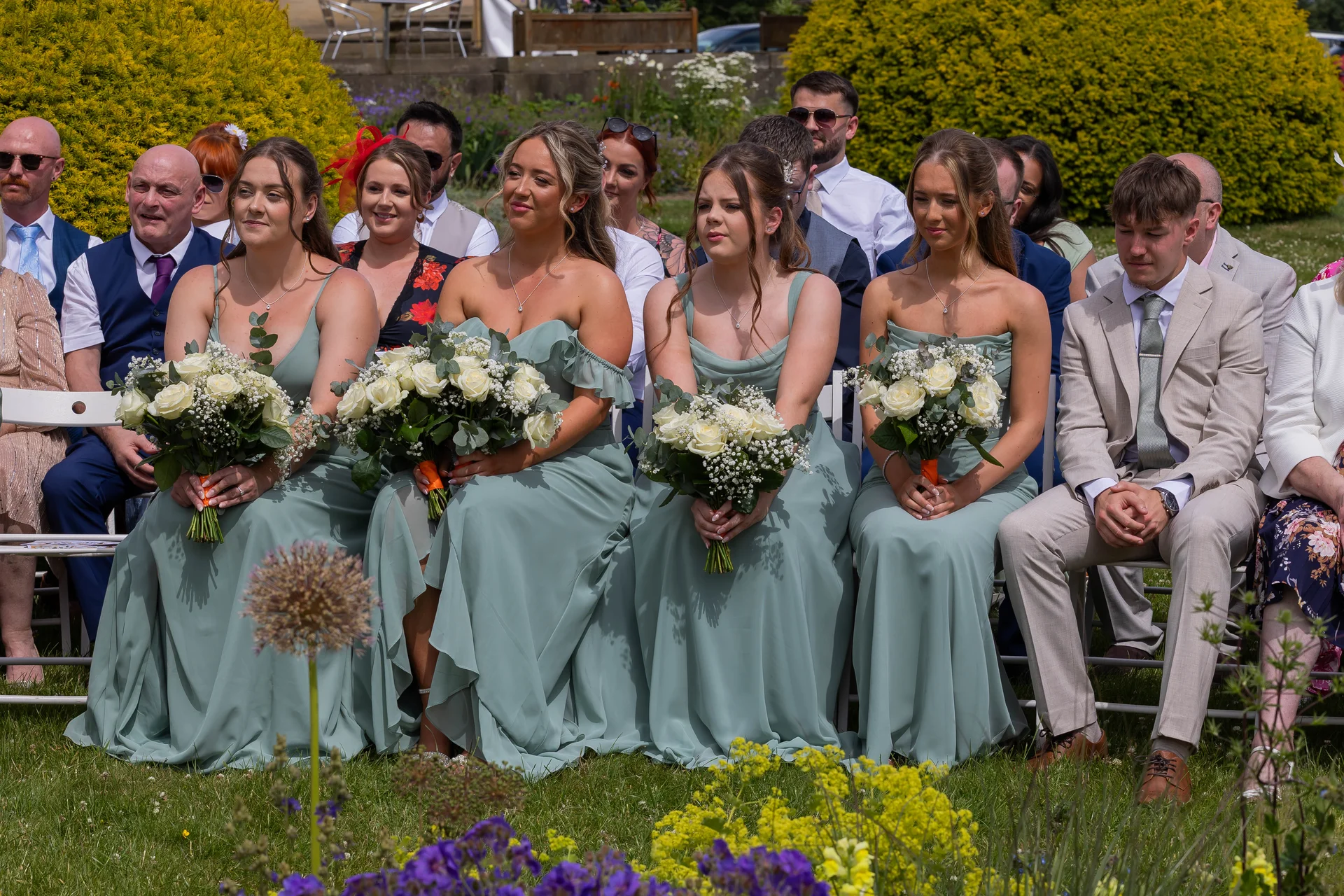 A row of bridesmaids wearing matching sage green dresses and holding white rose bouquets sits on white chairs during an outdoor wedding ceremony.