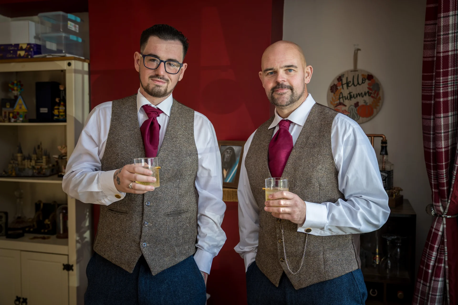 Two grooms in matching tweed waistcoats and burgundy ties smile as they hold glasses of whiskey during morning preparations.