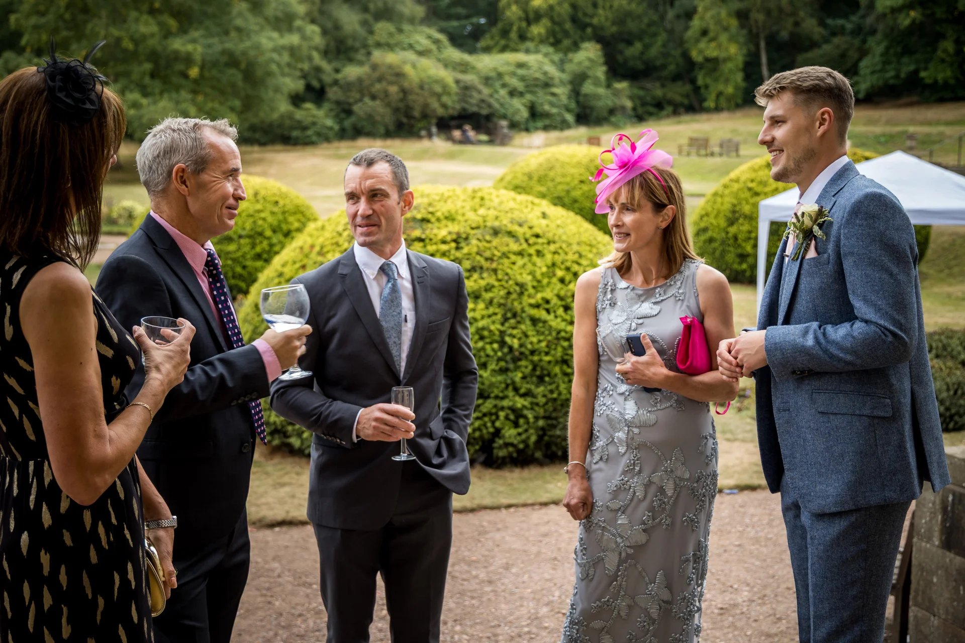 A group of well-dressed wedding guests mingling and chatting on a gravel path outdoors in the gardens during a drinks reception.