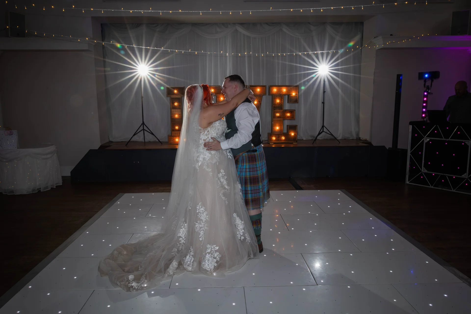 Newlyweds share their first dance together on an illuminated LED dance floor in front of glowing marquee letters.