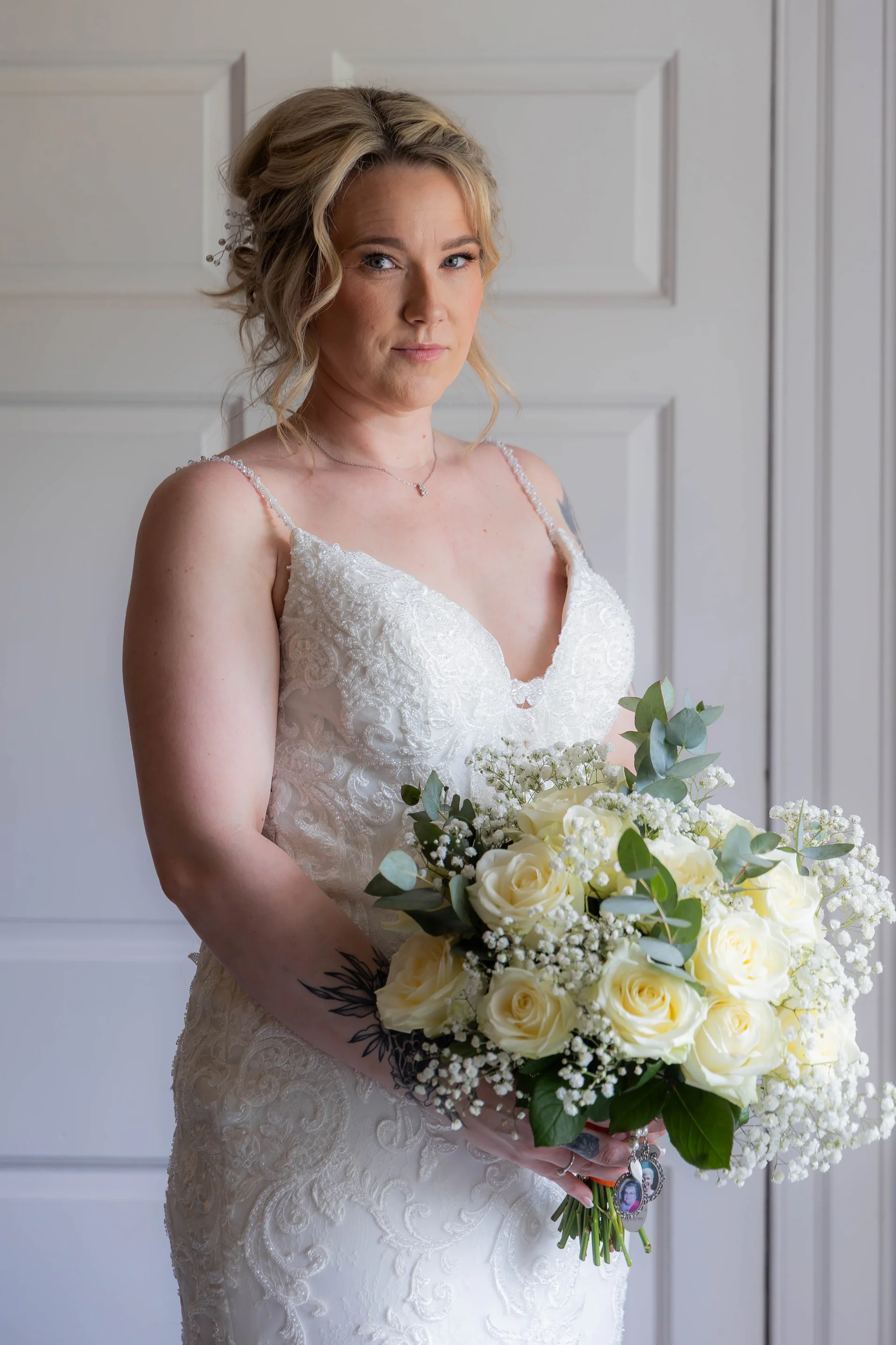 A stunning bride stands in a doorway, holding a lush hand-tied bouquet of white roses, baby's breath, and eucalyptus.