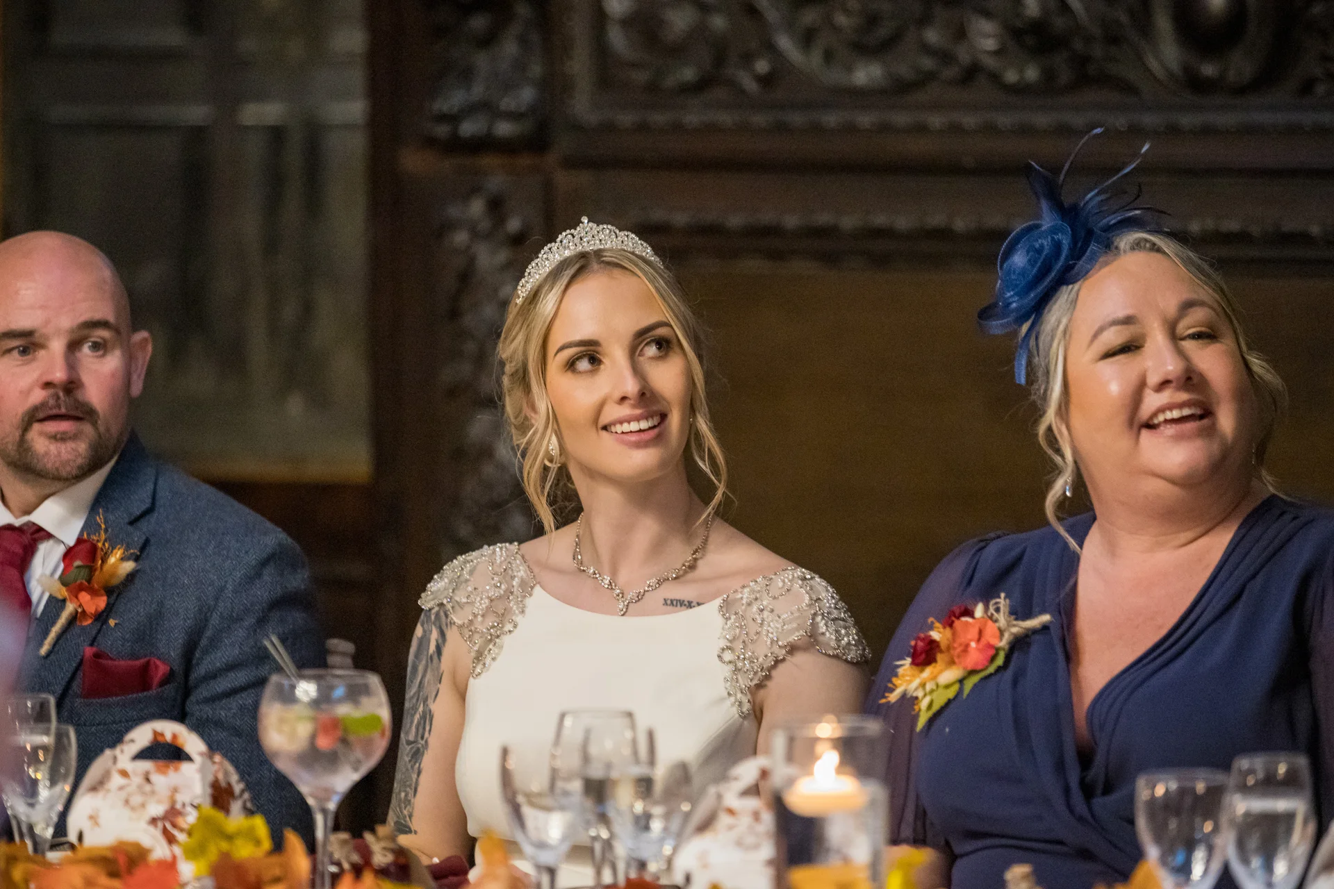 A smiling bride wearing a tiara and beaded gown sits alongside guests during wedding reception speeches in a wood-panelled room.