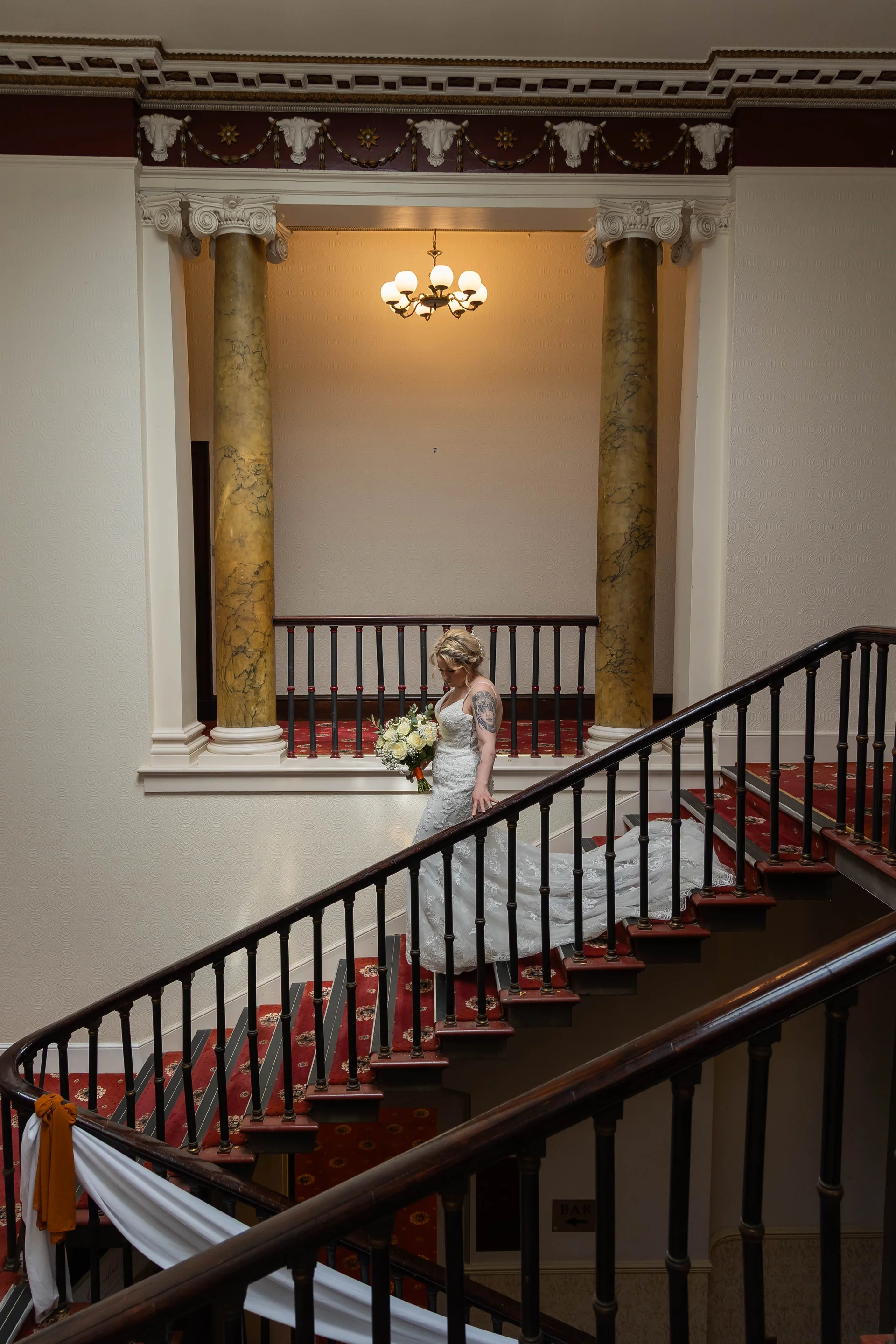 A stunning bride descends a grand carpeted staircase inside a classic country house holding a bouquet of white roses.