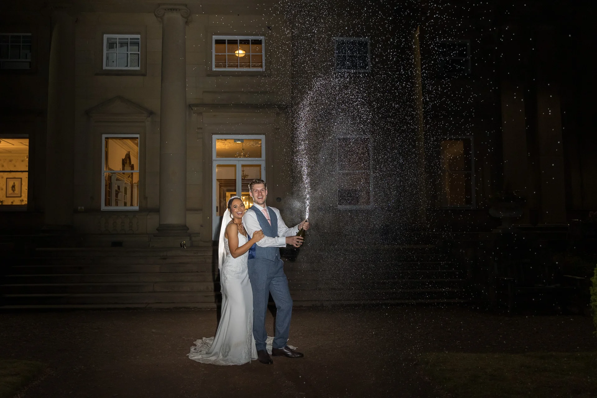 A newlywed couple laugh together in front of a grand illuminated country house at night while spraying a bottle of champagne.