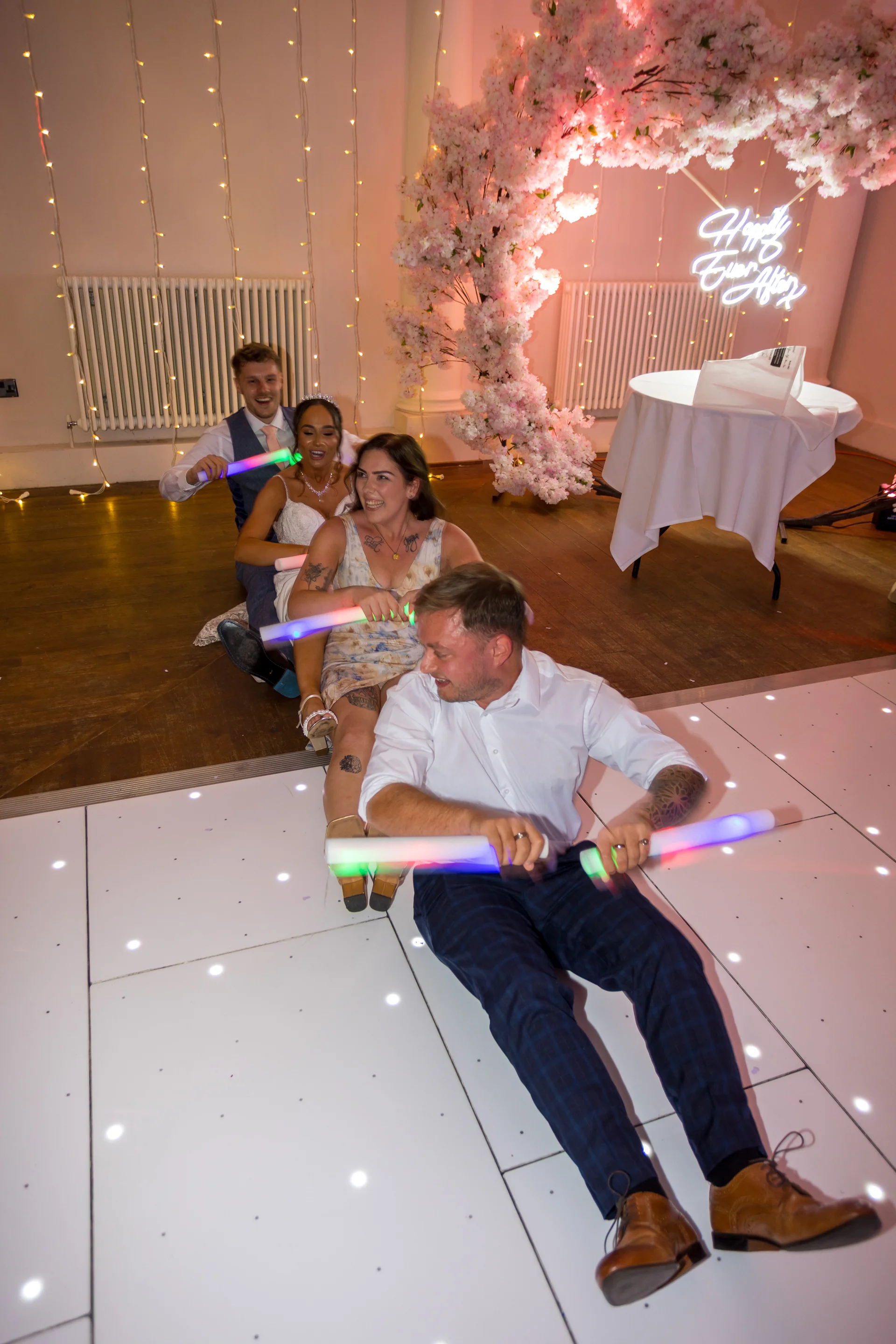 A bride, groom, and wedding guests sit in a row on a lit dance floor, pretending to row a boat with glowing foam sticks beneath a floral arch.