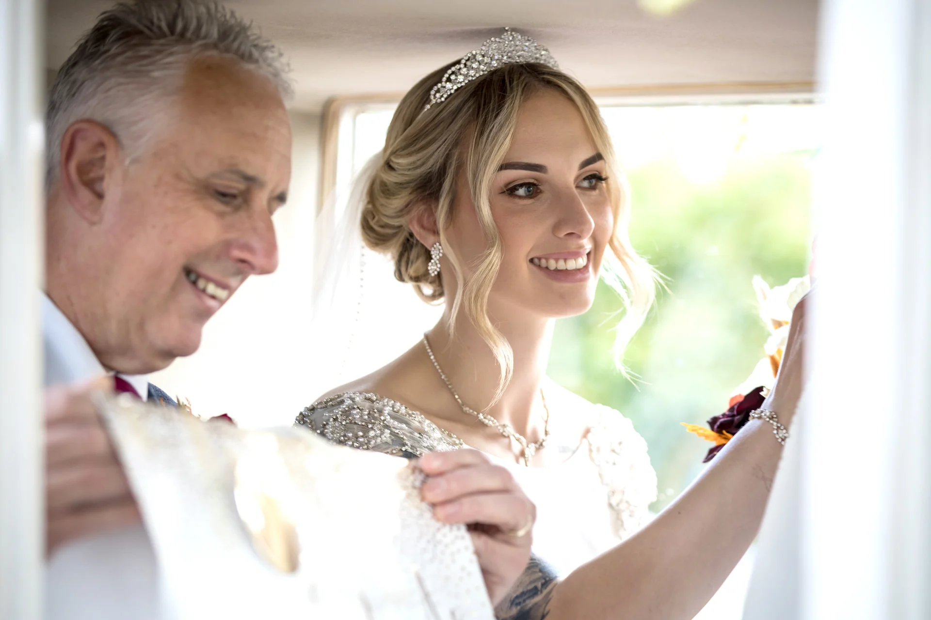 A smiling bride wearing a crystal tiara and beaded gown looks out of a wedding car window alongside her father.