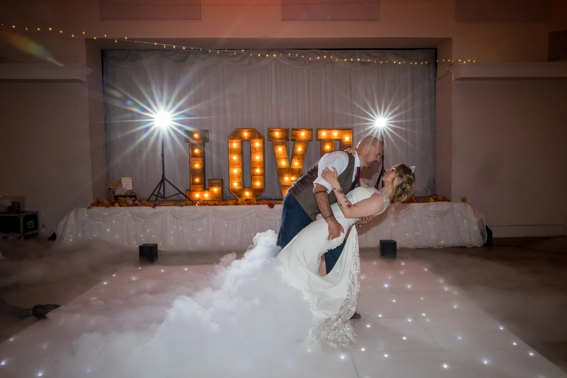 A bride and groom share a romantic dip kiss on a lit dance floor surrounded by low-lying smoke, with a illuminated love sign in the background.