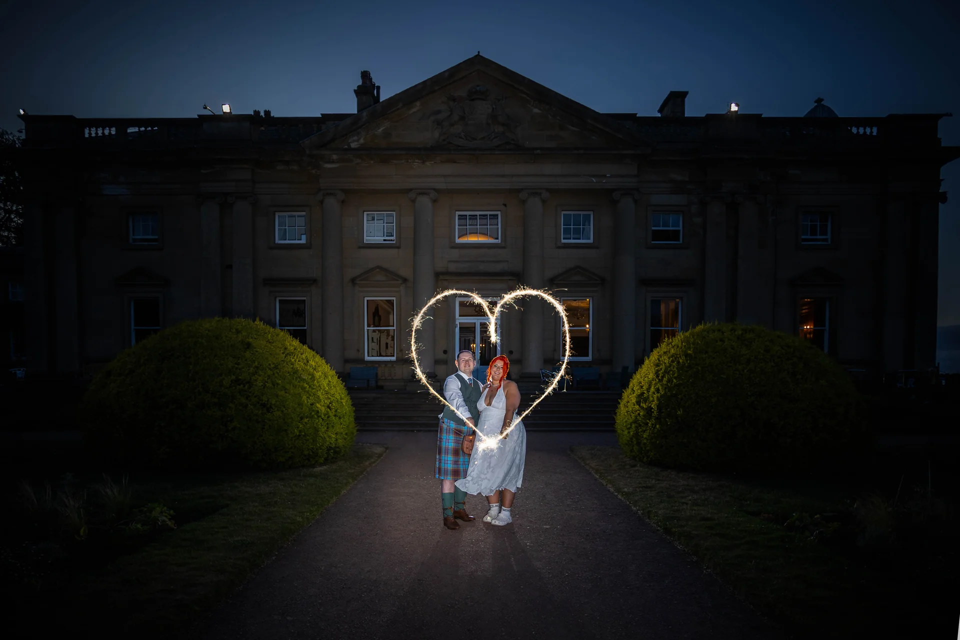 A newlywed couple poses in front of a grand illuminated country house at night while a sparkler is drawn into a heart shape around them.