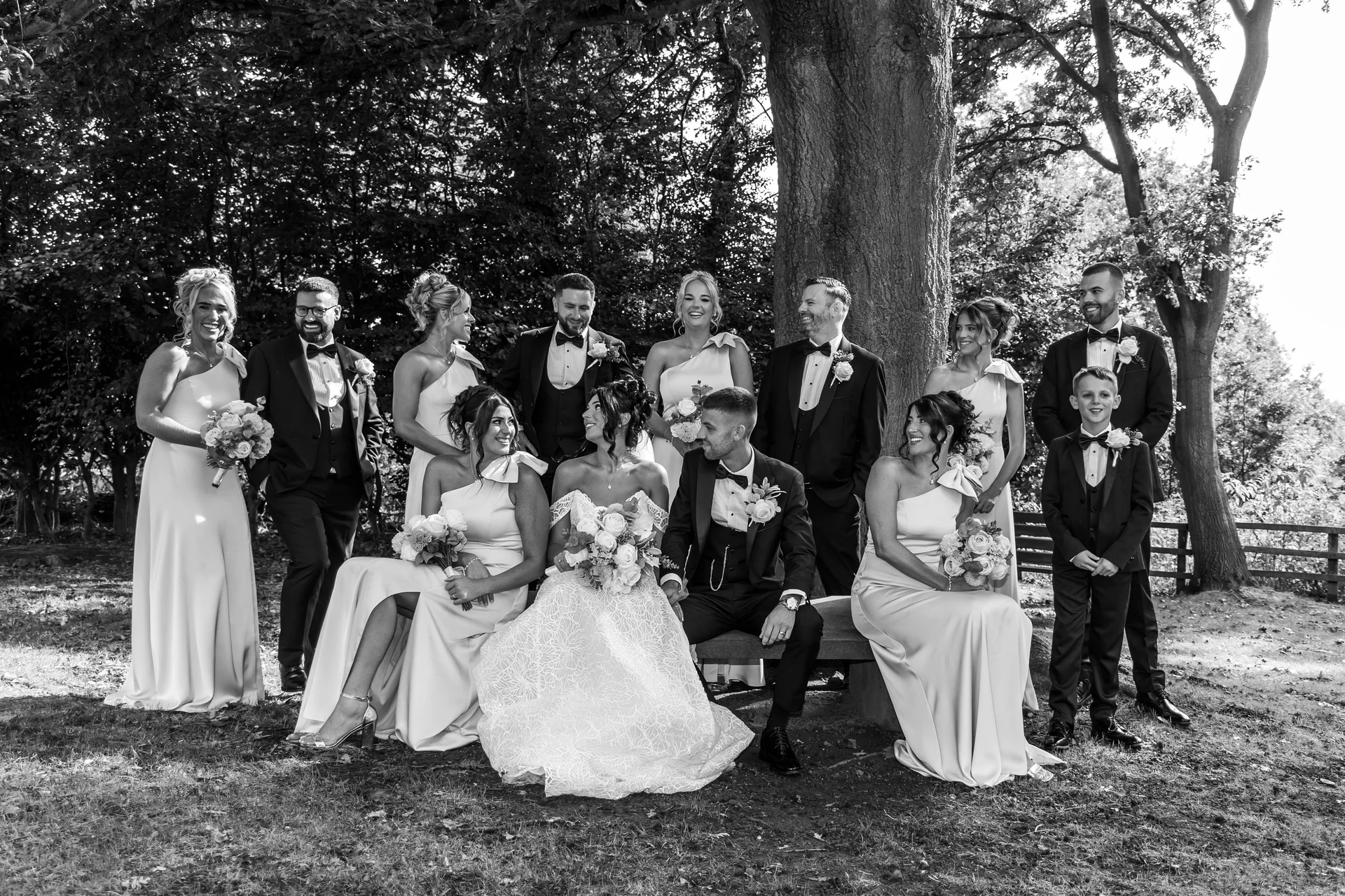 A bridal party poses outdoors in a woodland setting beneath a large tree, with the bride and groom seated on a stone bench surrounded by smiling bridesmaids in matching one-shoulder dresses and groomsmen in black tuxedos.