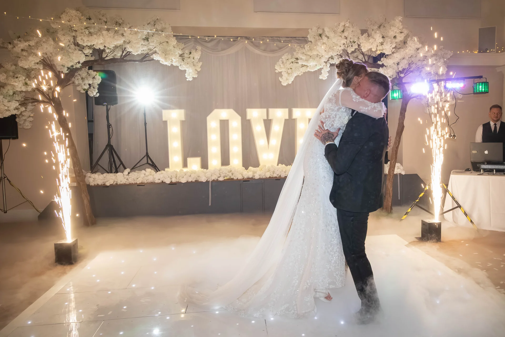 The newlyweds share an embrace on a illuminated dance floor surrounded by low-lying smoke, cold sparklers, and large decorative lit letters.