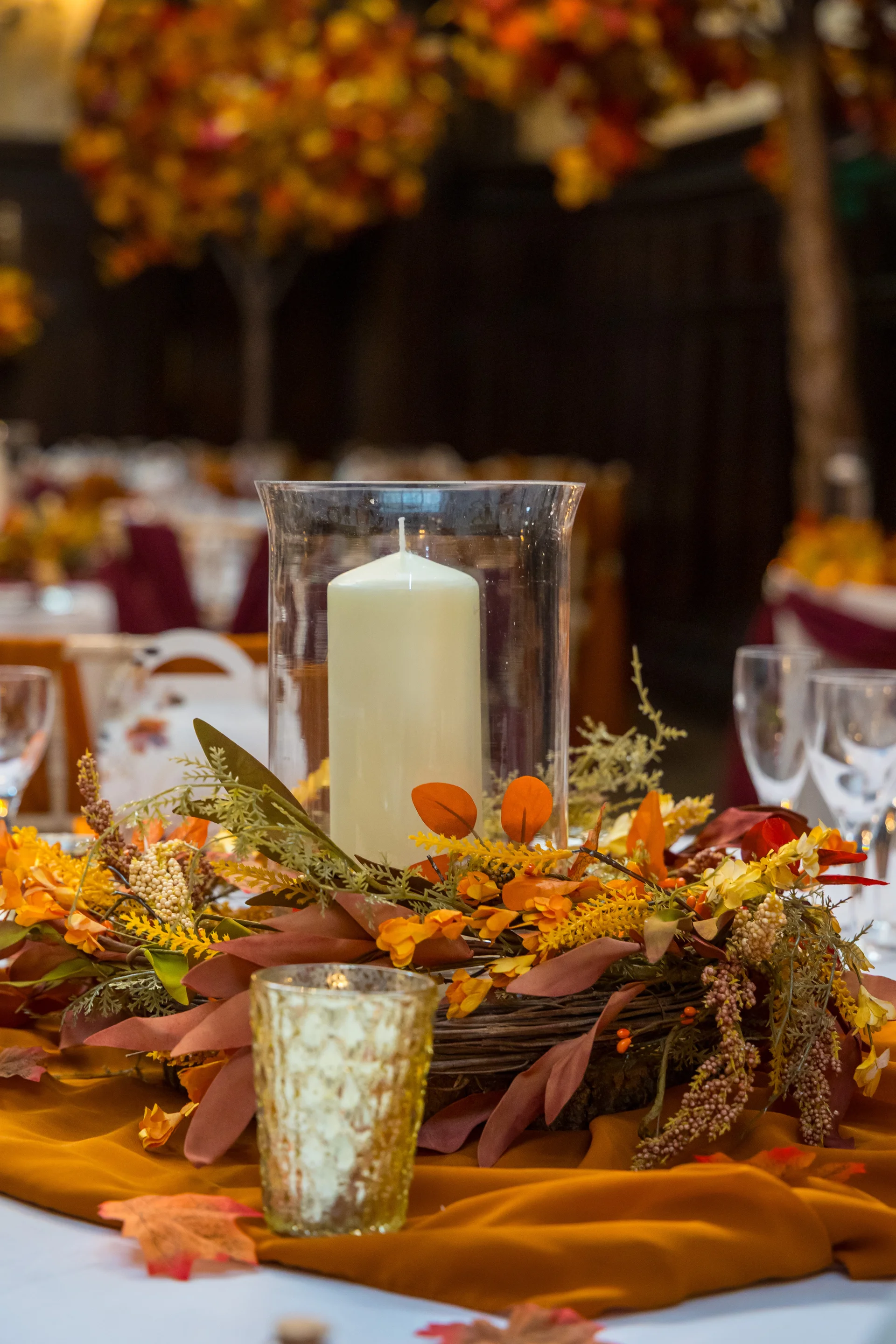 An autumnal wedding tablescape features a pillar candle in a glass hurricane vase resting inside a rich floral wreath of orange leaves and berries.