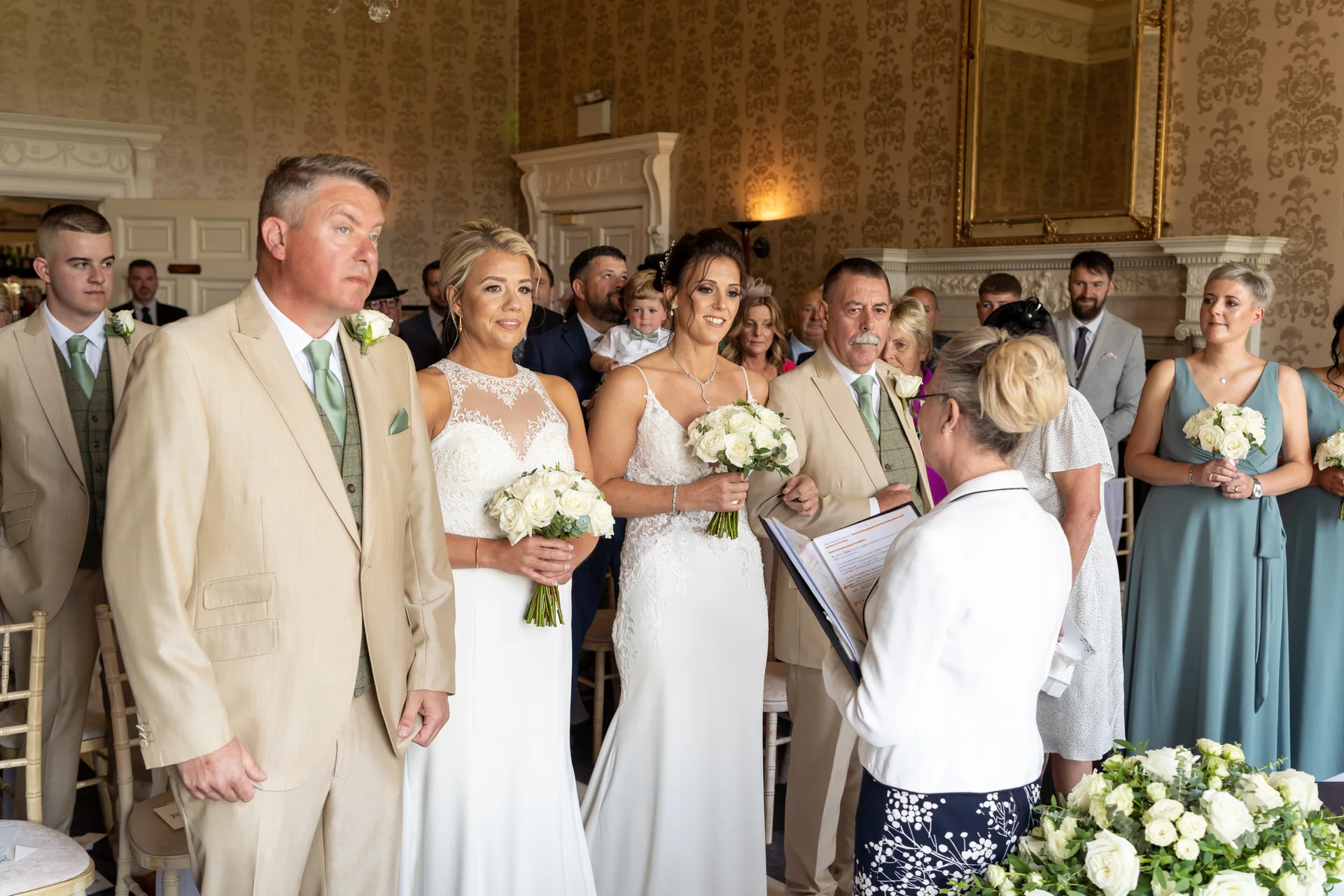 Two brides stand together holding bouquets of white roses during a civil ceremony in a grand panelled room, surrounded by family and wedding party members.