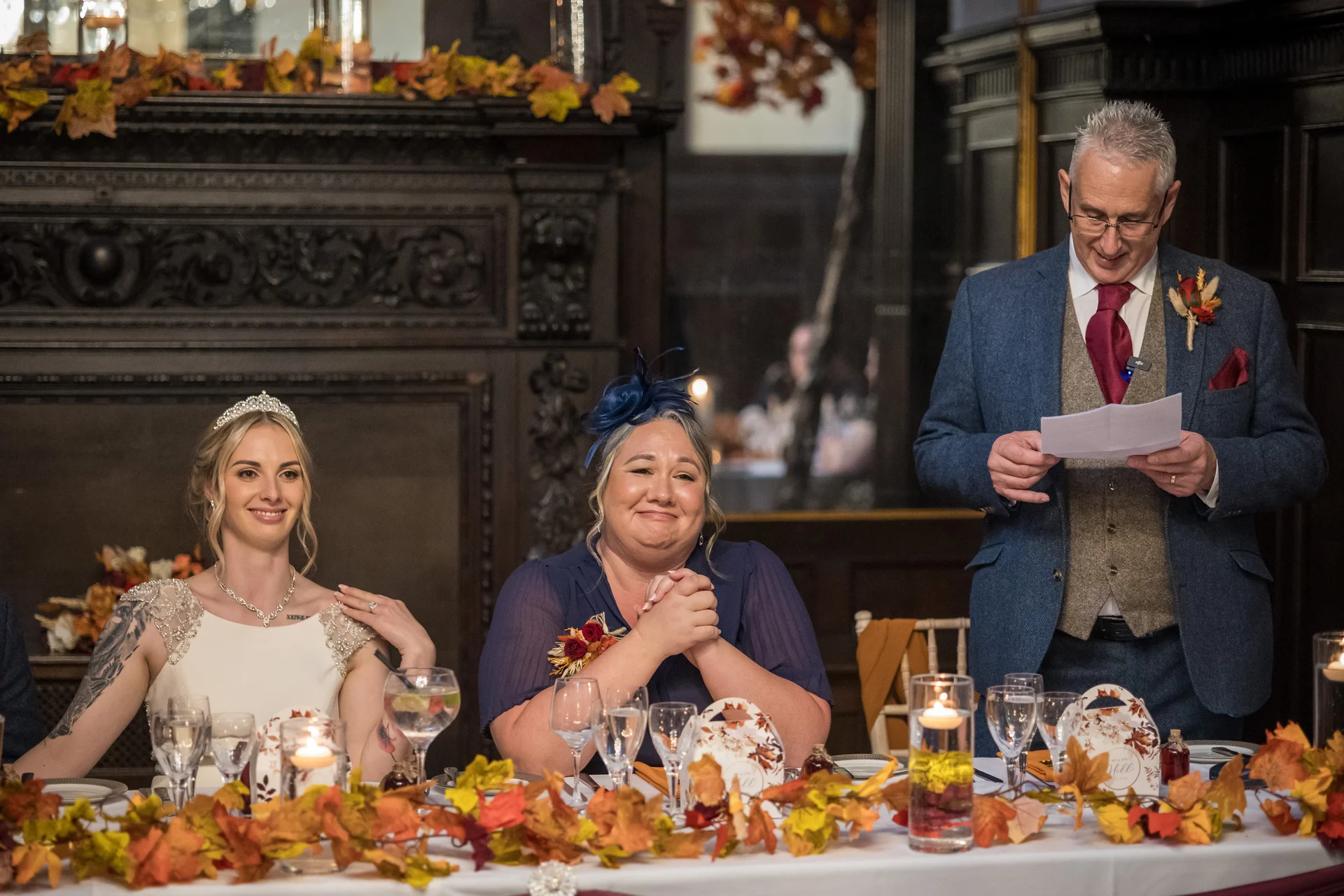 A bride and a guest sit smiling at the top table while a man in a tweed waistcoat and suit jacket delivers a speech during an autumnal wedding reception.