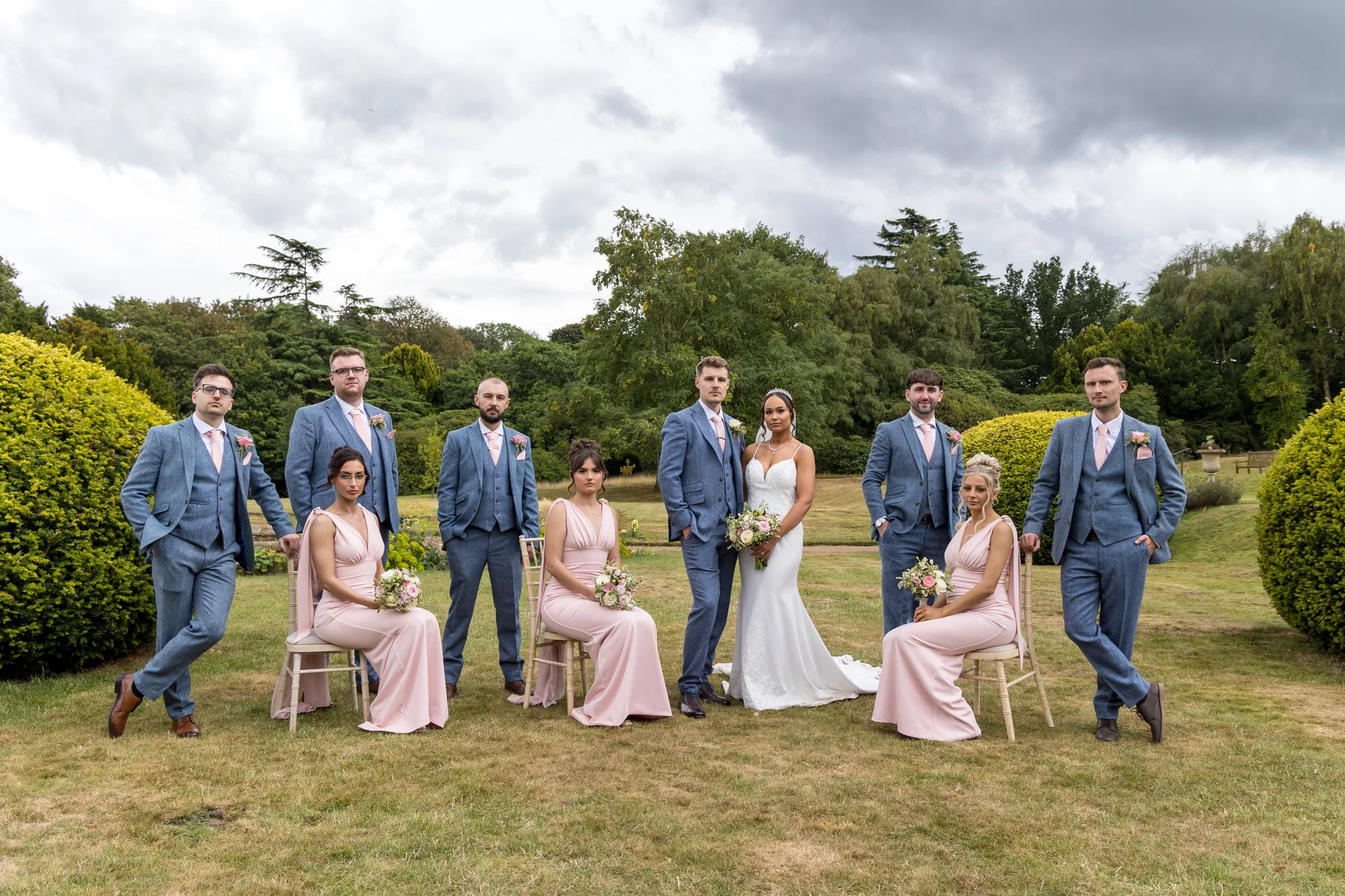 A bridal party poses outdoors in a manicured garden on an overcast day, featuring the bride in a white gown flanked by groomsmen in blue tweed suits and seated bridesmaids in pale pink dresses.