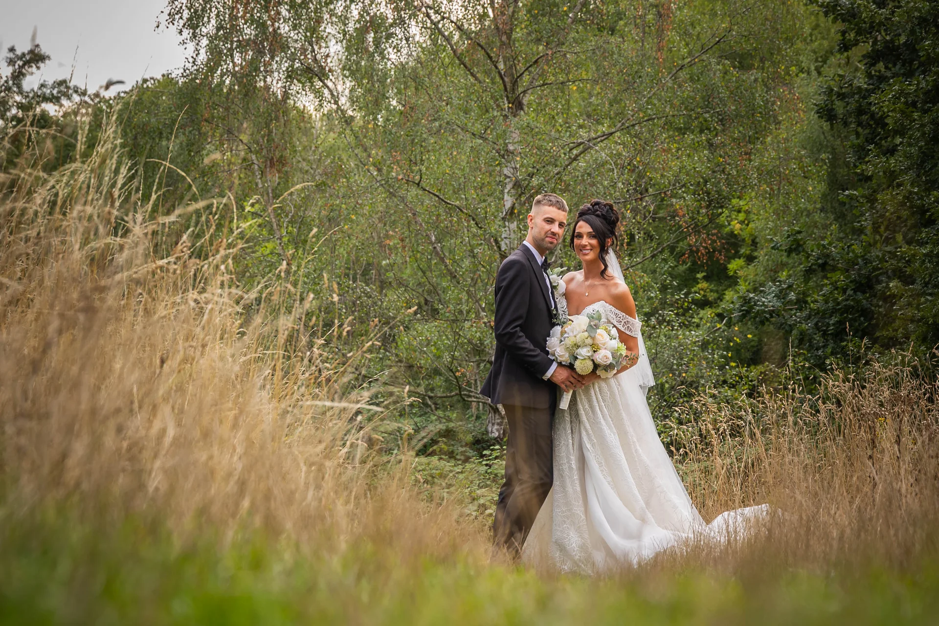 A newlywed couple poses in a lush woodland setting, surrounded by tall wild grass and mature trees.