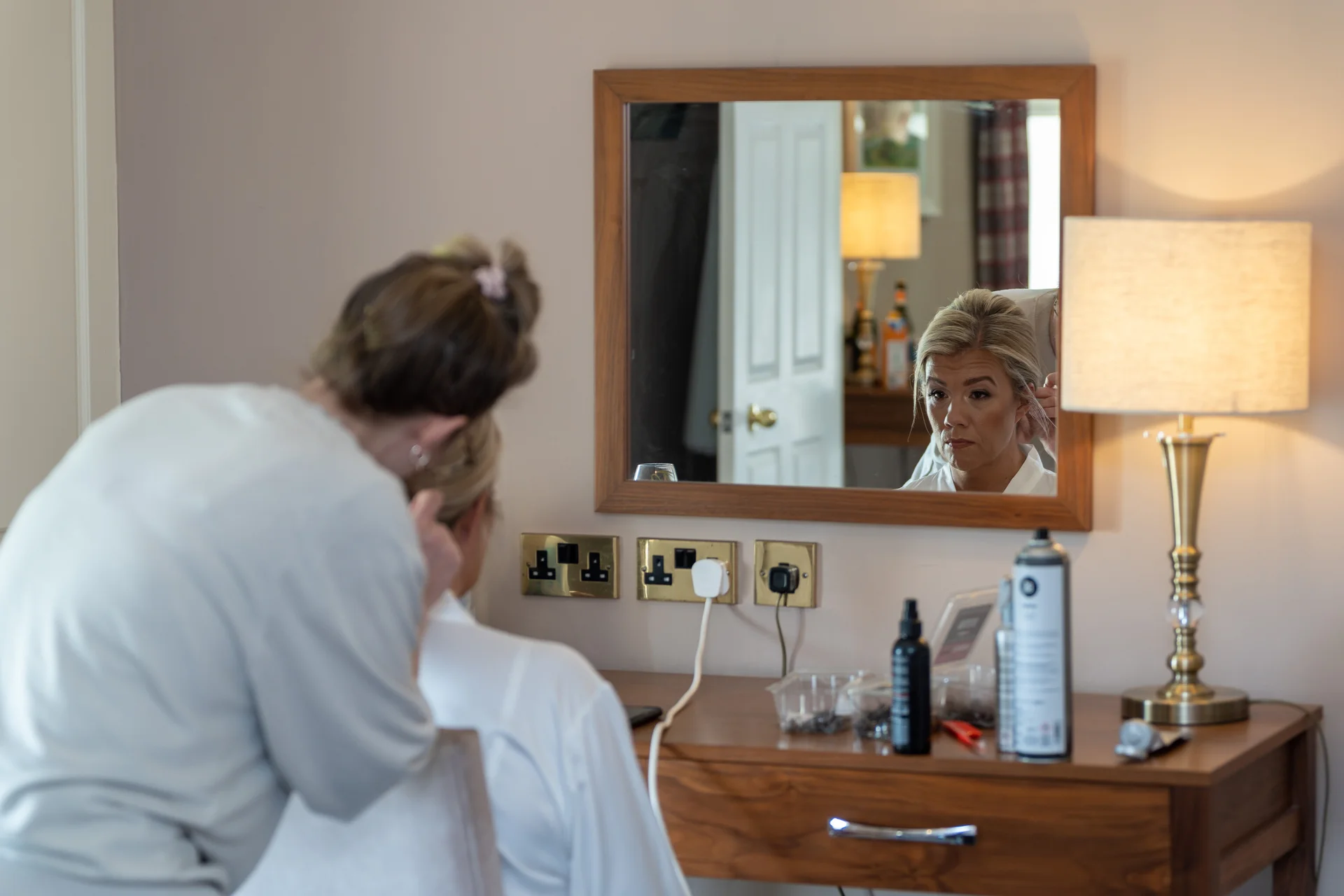 A bride sits in front of a dressing table mirror while having her hair styled during wedding morning preparations.