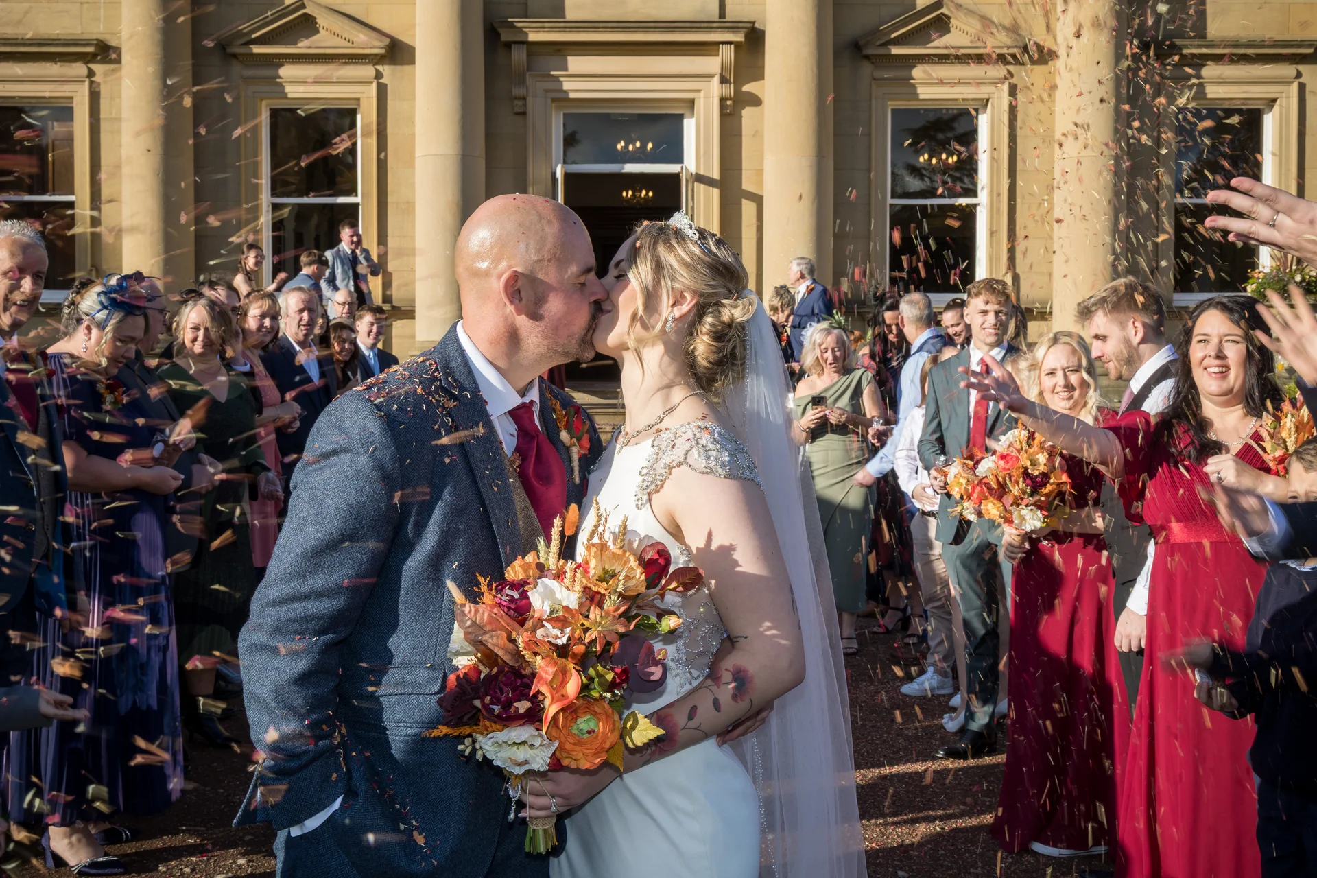 Newlyweds share a romantic kiss outside a grand country house as cheering wedding guests shower them in colourful confetti during an autumn celebration.