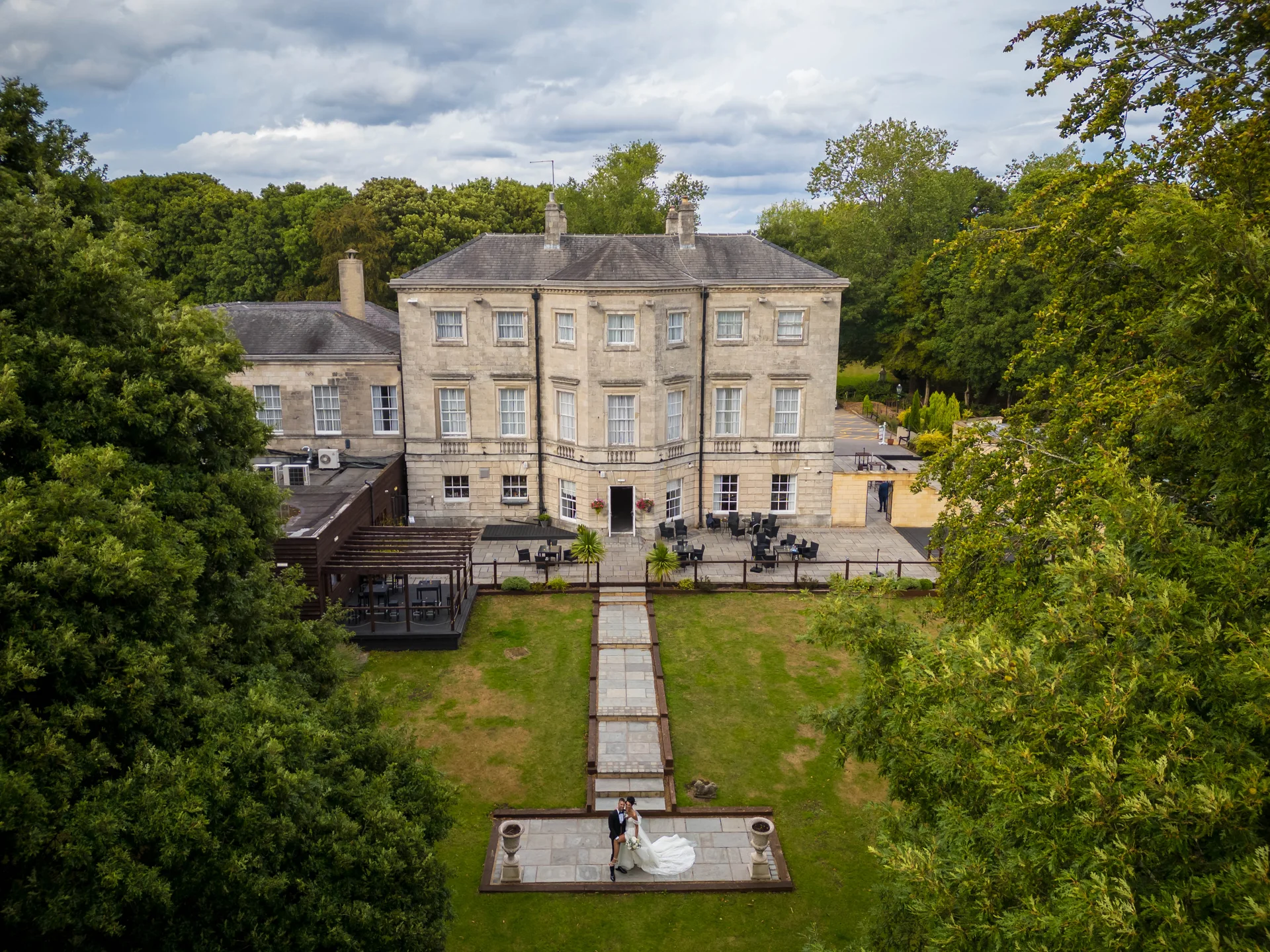 An aerial drone shot captures a newlywed couple standing on a stone patio in front of a grand country house surrounded by lush trees and manicured lawns.