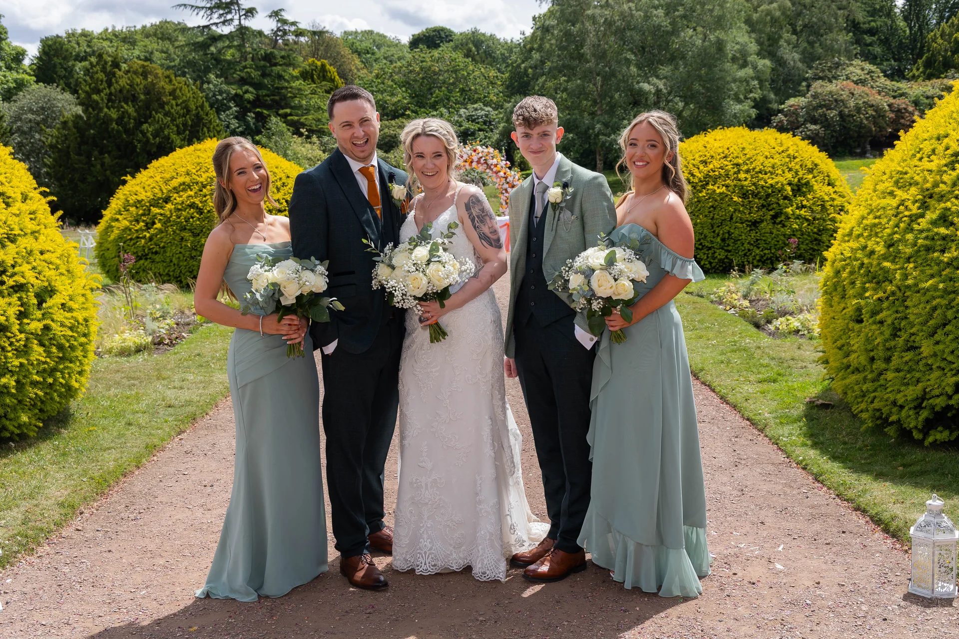 A newlywed couple poses outdoors in a lush garden pathway with two bridesmaids and a groomsman, all holding white rose bouquets and laughing happily.