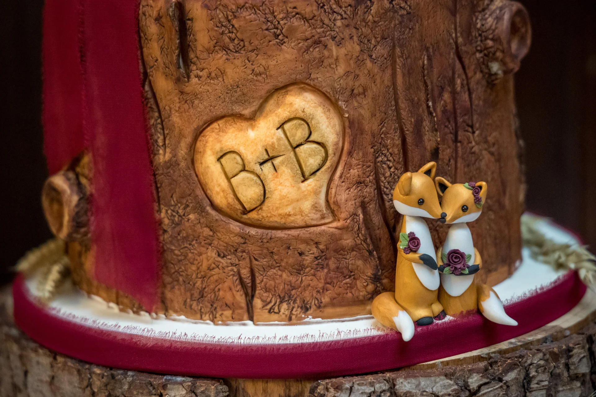 A whimsical woodland-themed wedding cake is decorated with realistic tree bark fondant, a carved heart showing initials, and two adorable handmade fox cake toppers.