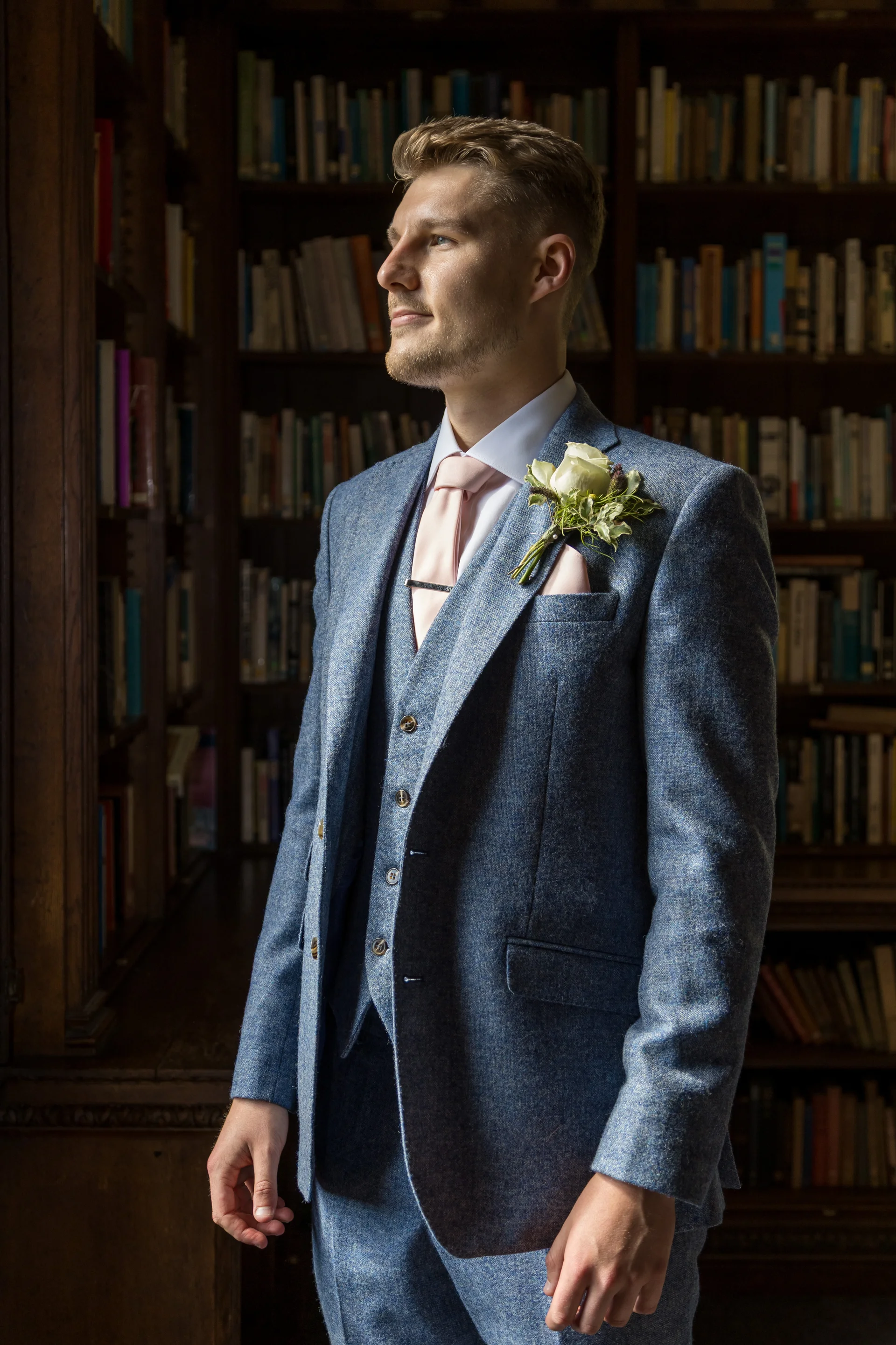 A handsome groom stands proudly in a library wearing a blue tweed three-piece suit adorned with a pink tie and a delicate white floral buttonhole.