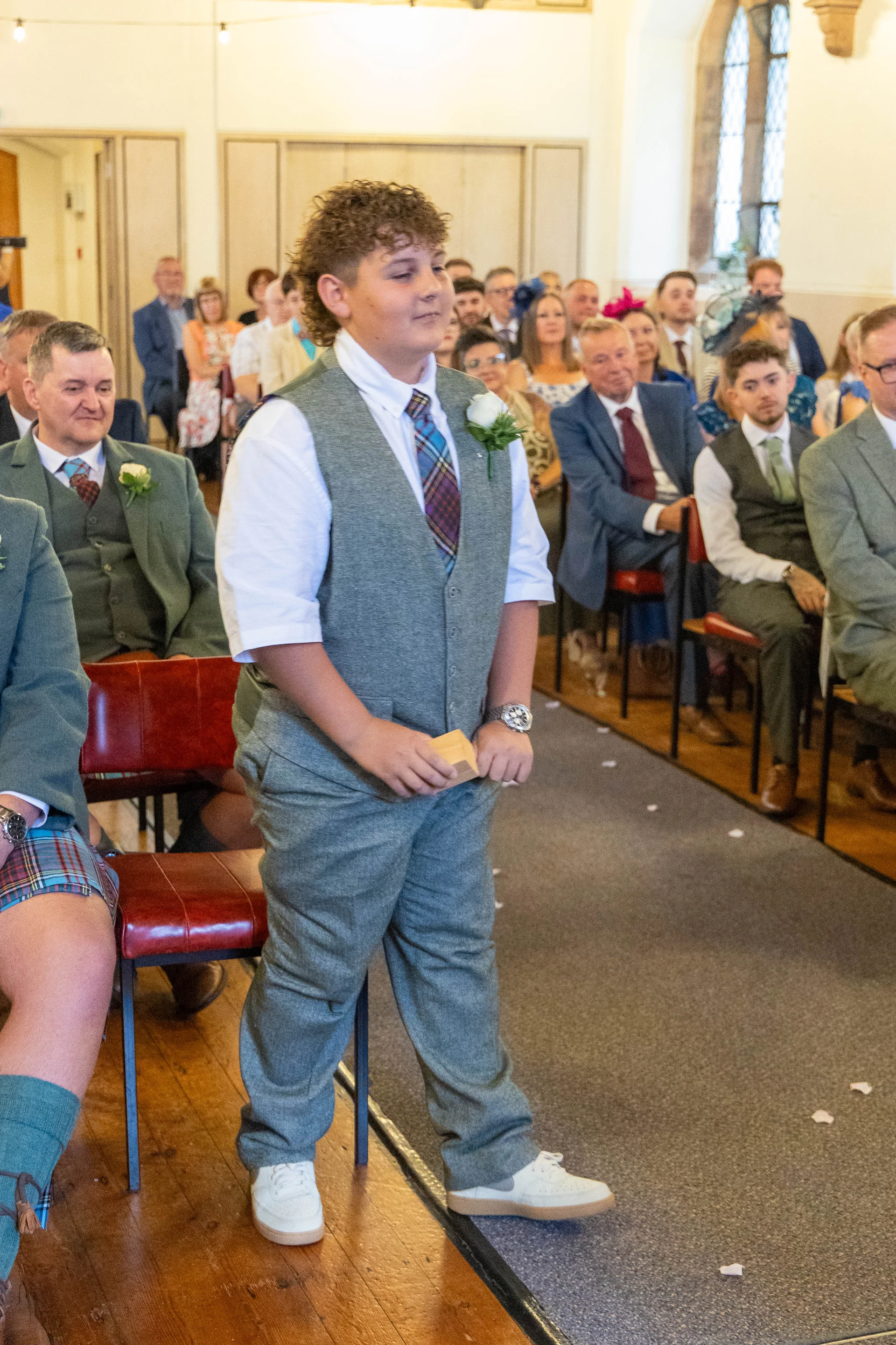 A young page boy wearing a grey tweed waistcoat, white shirt, and patterned tie stands at the front of a church during a wedding ceremony.