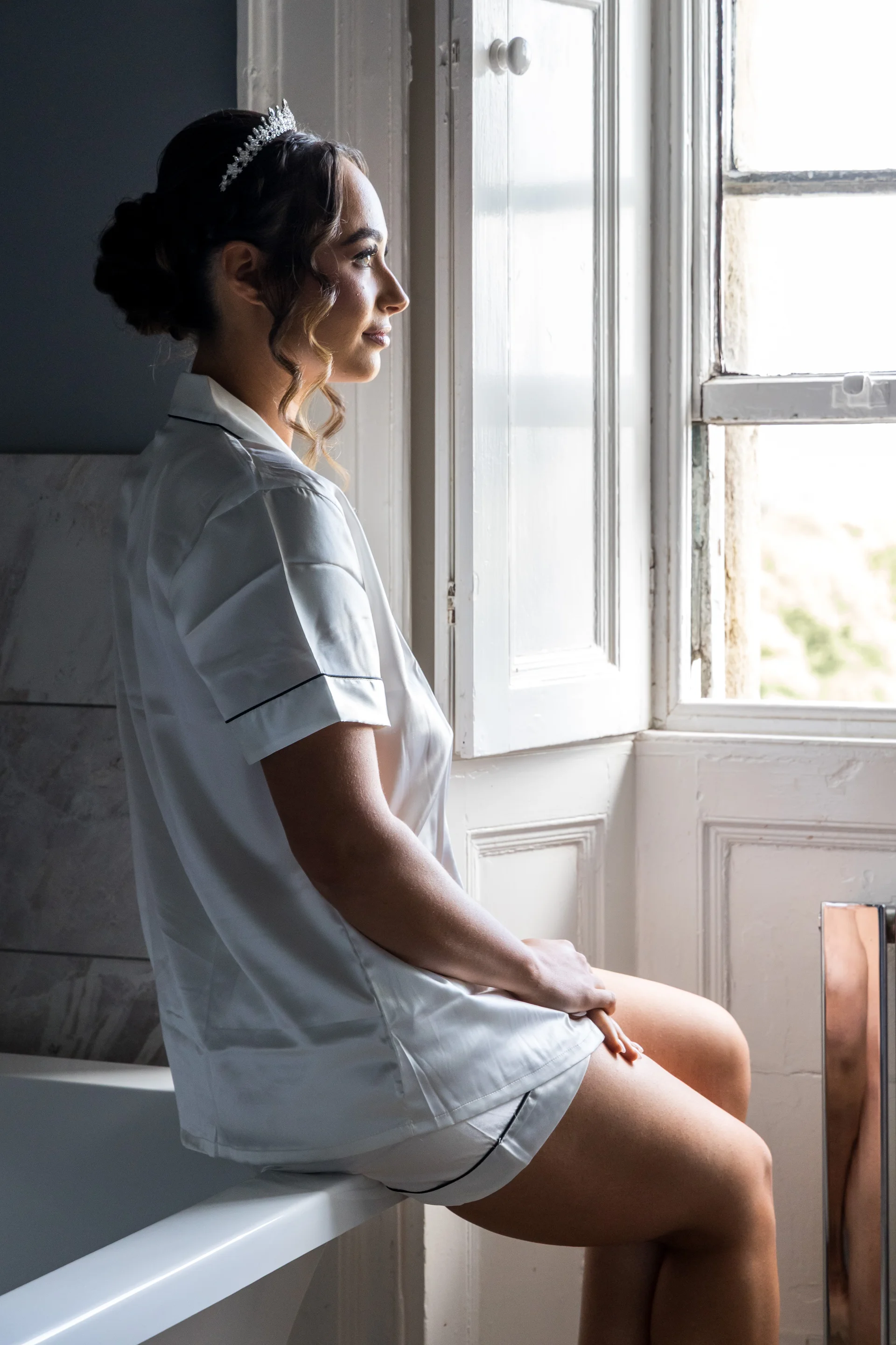 A beautiful bride sits by a bright window during wedding morning preparations, wearing matching white silk pyjamas and a sparkling tiara in her updo.