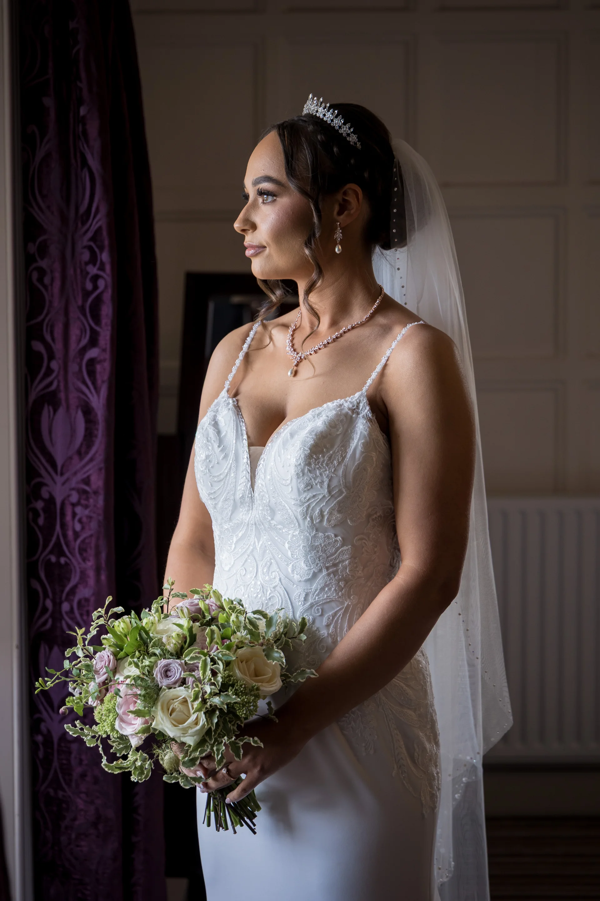 A glowing bride stands by a window in natural light, wearing a lace wedding dress, a crystal tiara, and holding a hand-tied floral bouquet.