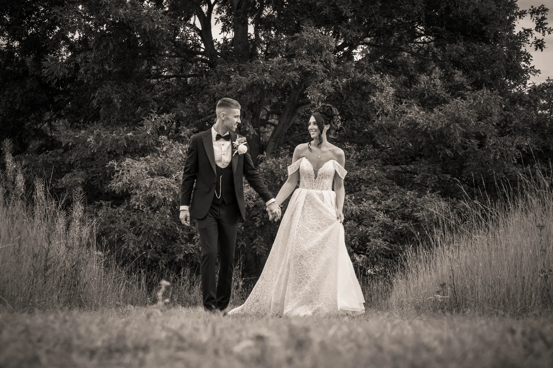 Newlyweds lovingly gaze at each other while walking hand in hand through tall grass in front of a large leafy tree.