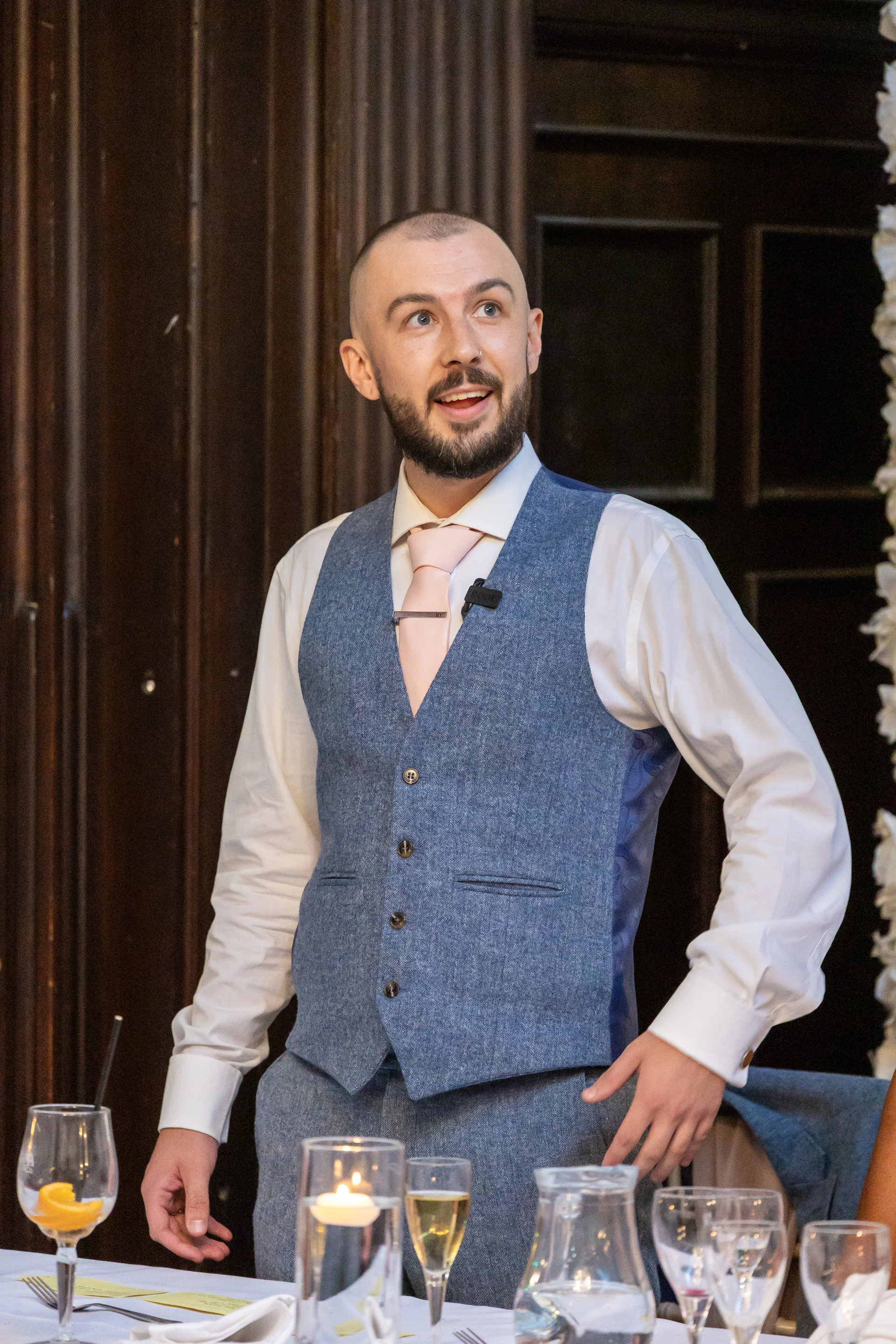 A smiling groom wearing a blue tweed waistcoat and pink tie delivers his speech during the wedding reception.