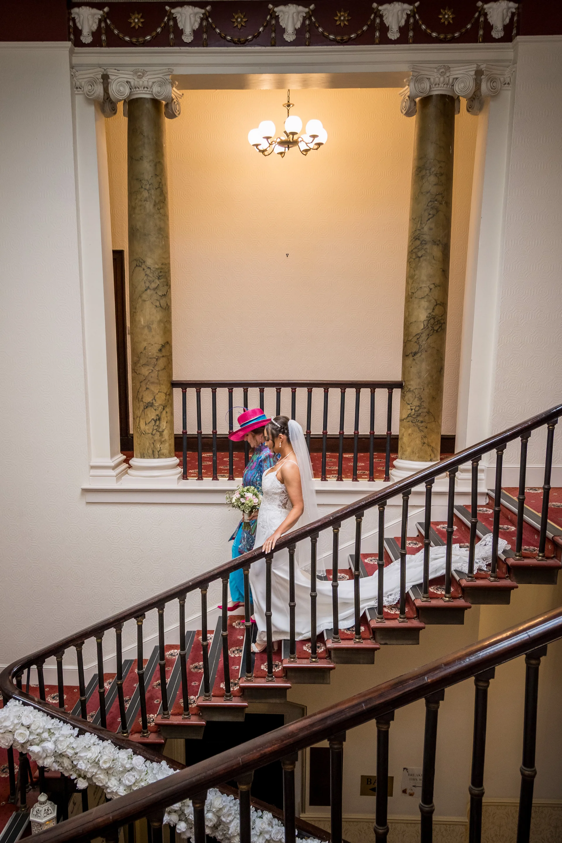 A bride in a classic white gown with a long train descends a grand staircase alongside the mother of the bride inside a historic venue.