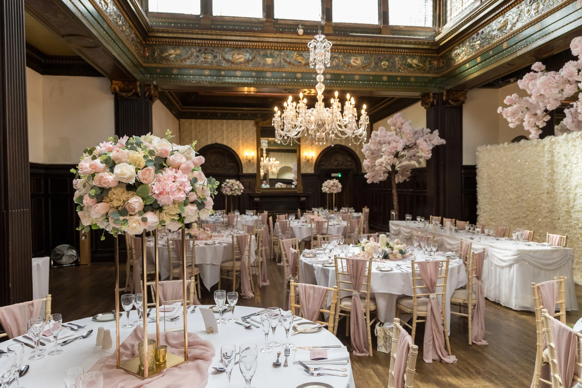 An opulent ballroom reception venue featuring elegantly dressed round tables with tall floral centrepieces, blush pink sashes, and a grand crystal chandelier overhead.