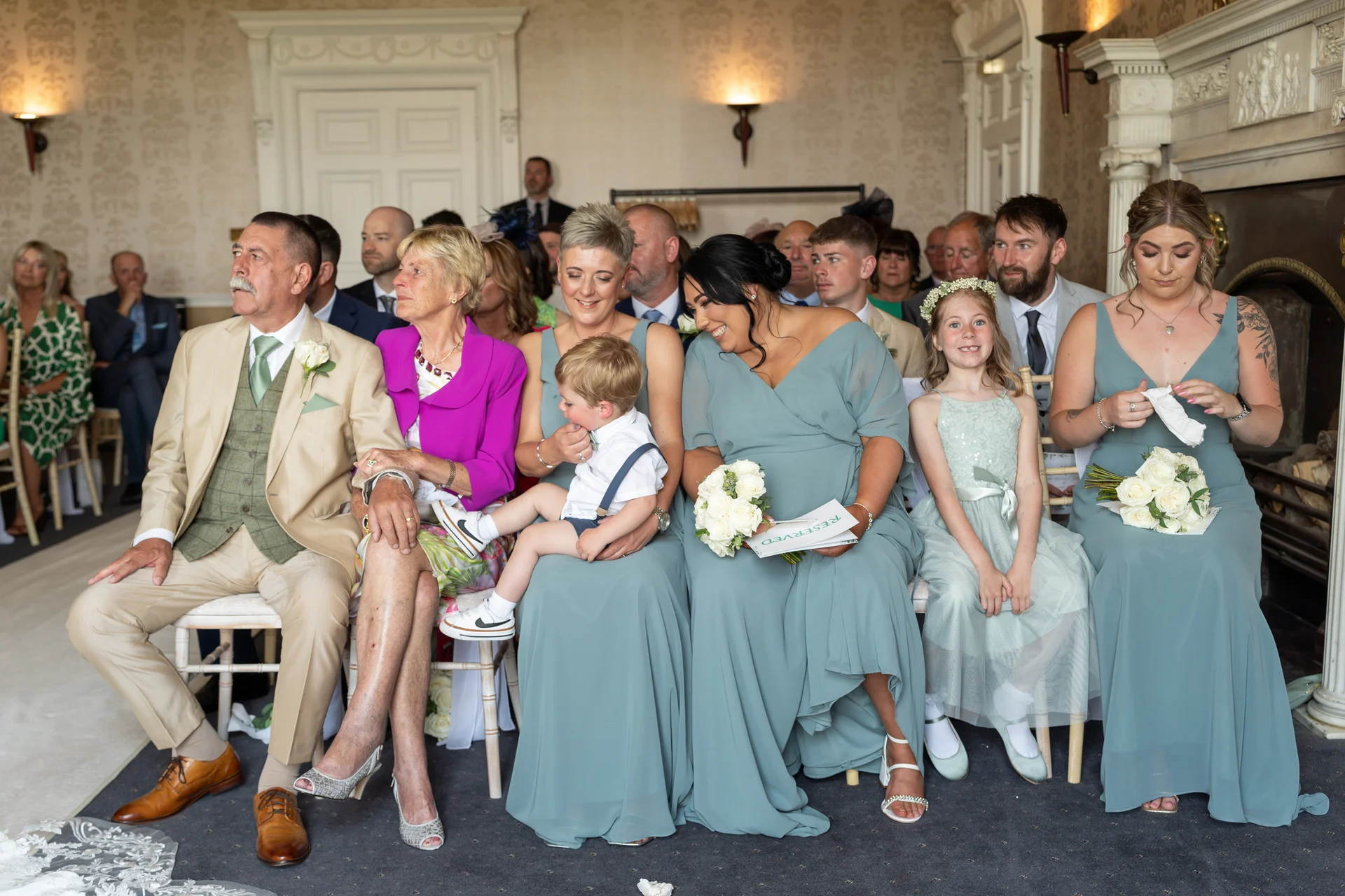 Wedding guests and bridal party members sit together indoors during a ceremony, with bridesmaids wearing sage green dresses and holding white rose bouquets.
