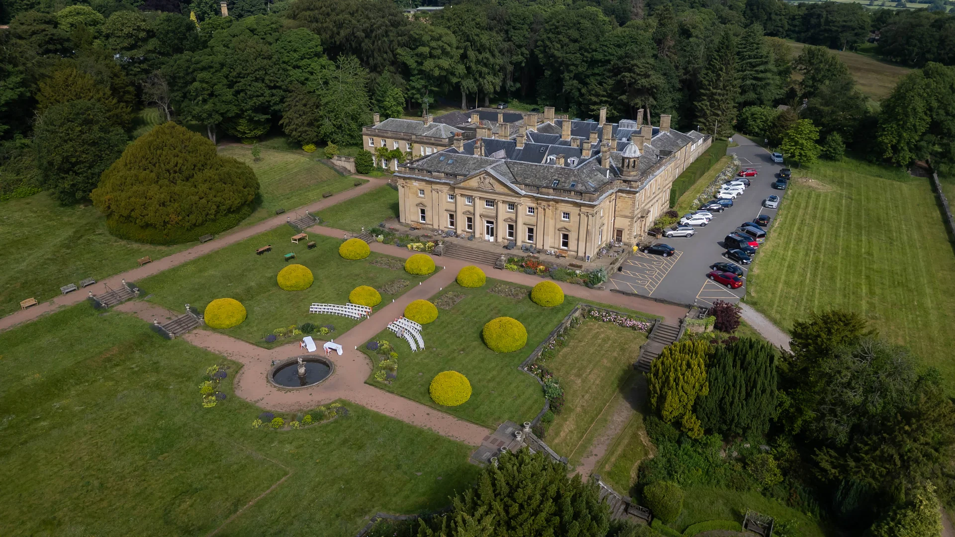 An aerial drone shot captures a grand stately home surrounded by lush green lawns, manicured gardens, and an outdoor ceremony setup with white chairs.