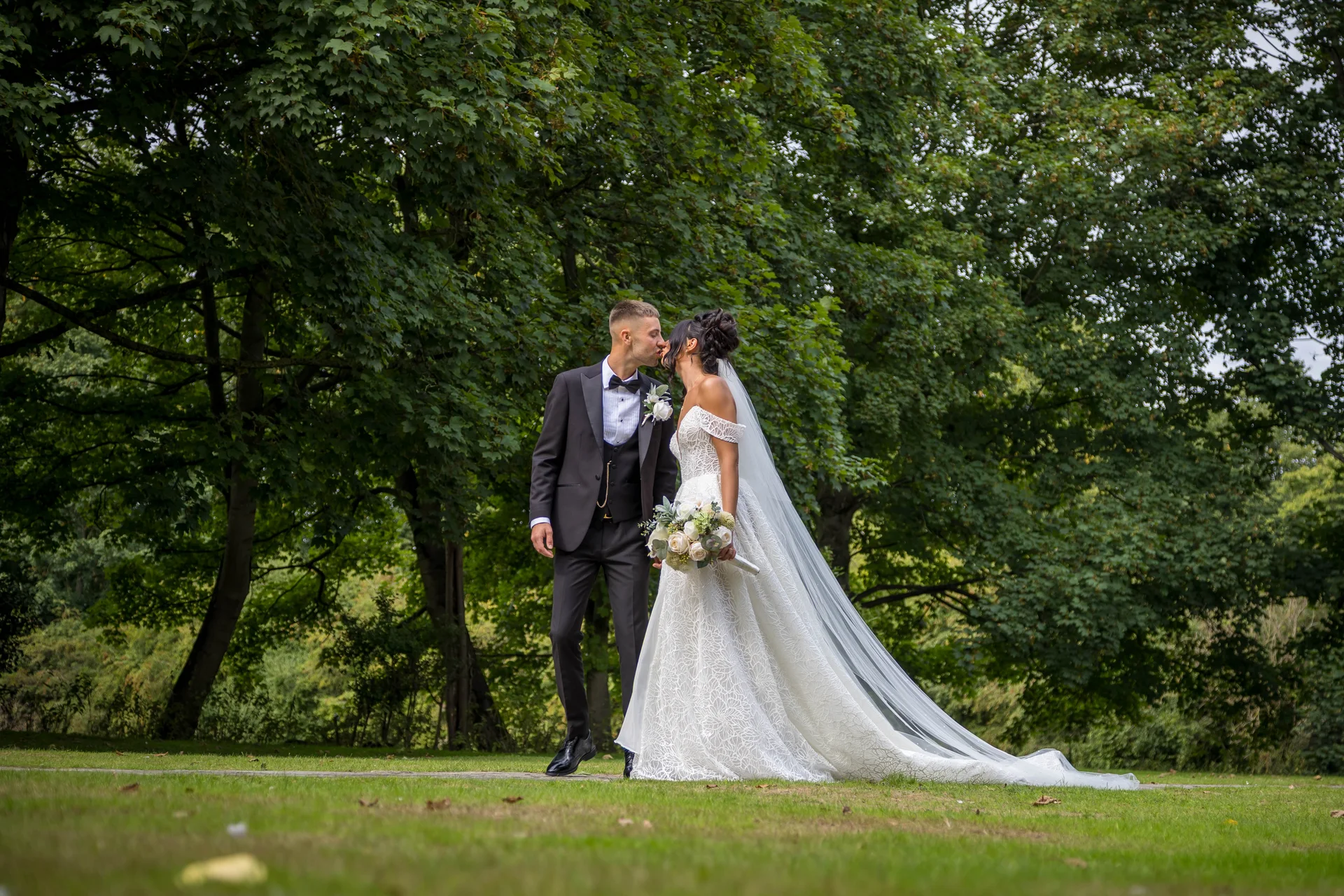 Newlyweds share a tender kiss outdoors against a lush green backdrop, with the bride wearing a detailed off-the-shoulder lace gown and a long veil.