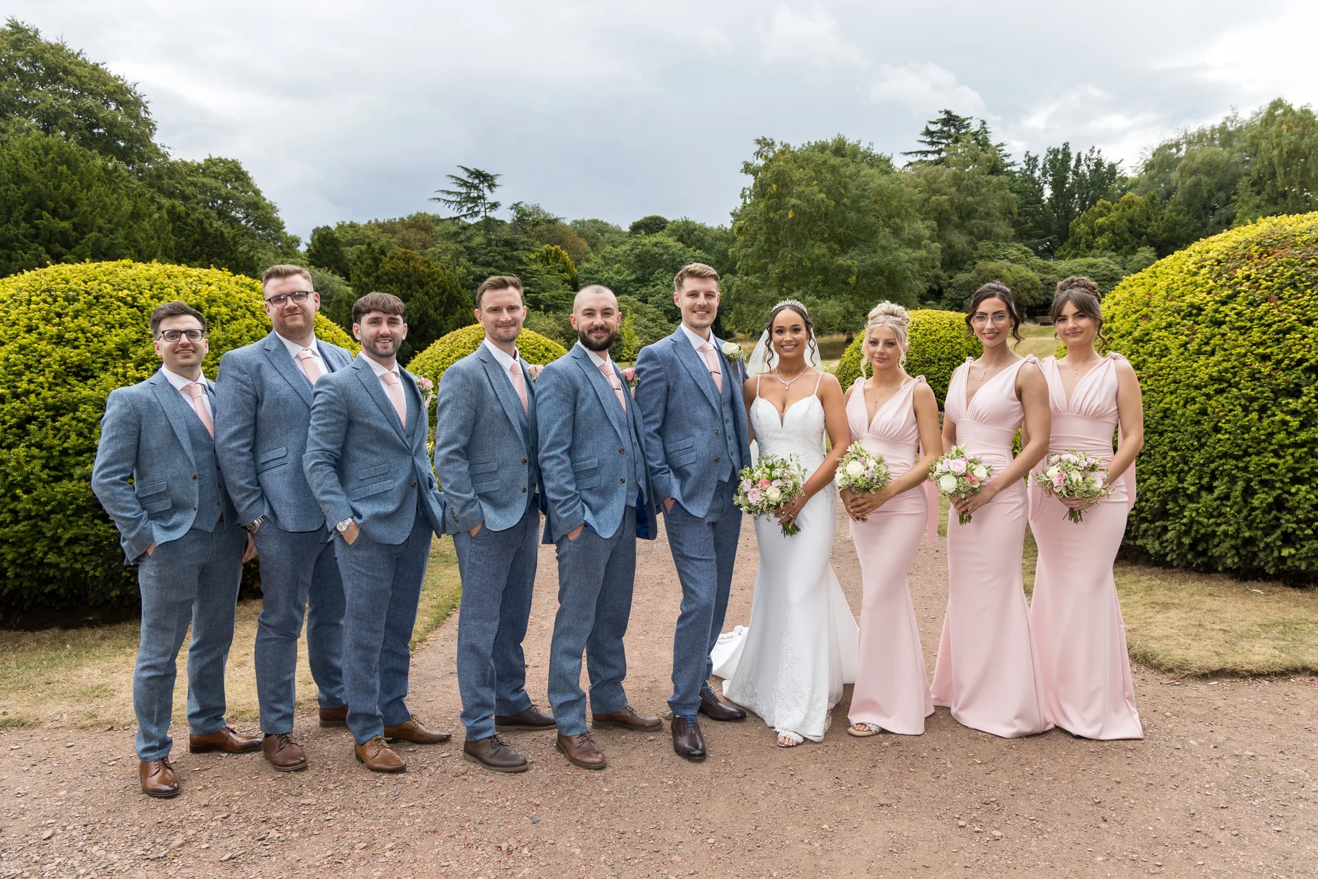 A bridal party of four groomsmen in blue suits and four bridesmaids in pink dresses poses outdoors with the bride and groom in front of lush green hedges.