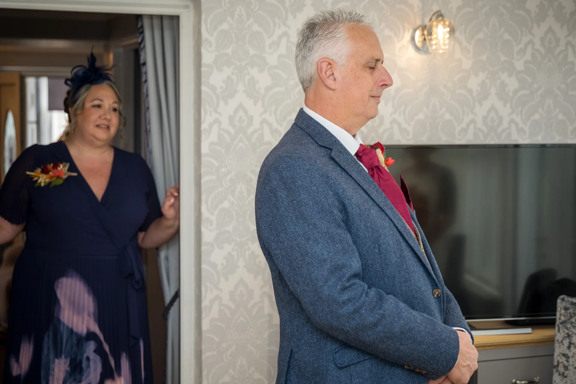 A mature groom in a textured blue suit waits calmly indoors while a woman wearing a navy dress and a fascinator watches from an adjoining doorway.