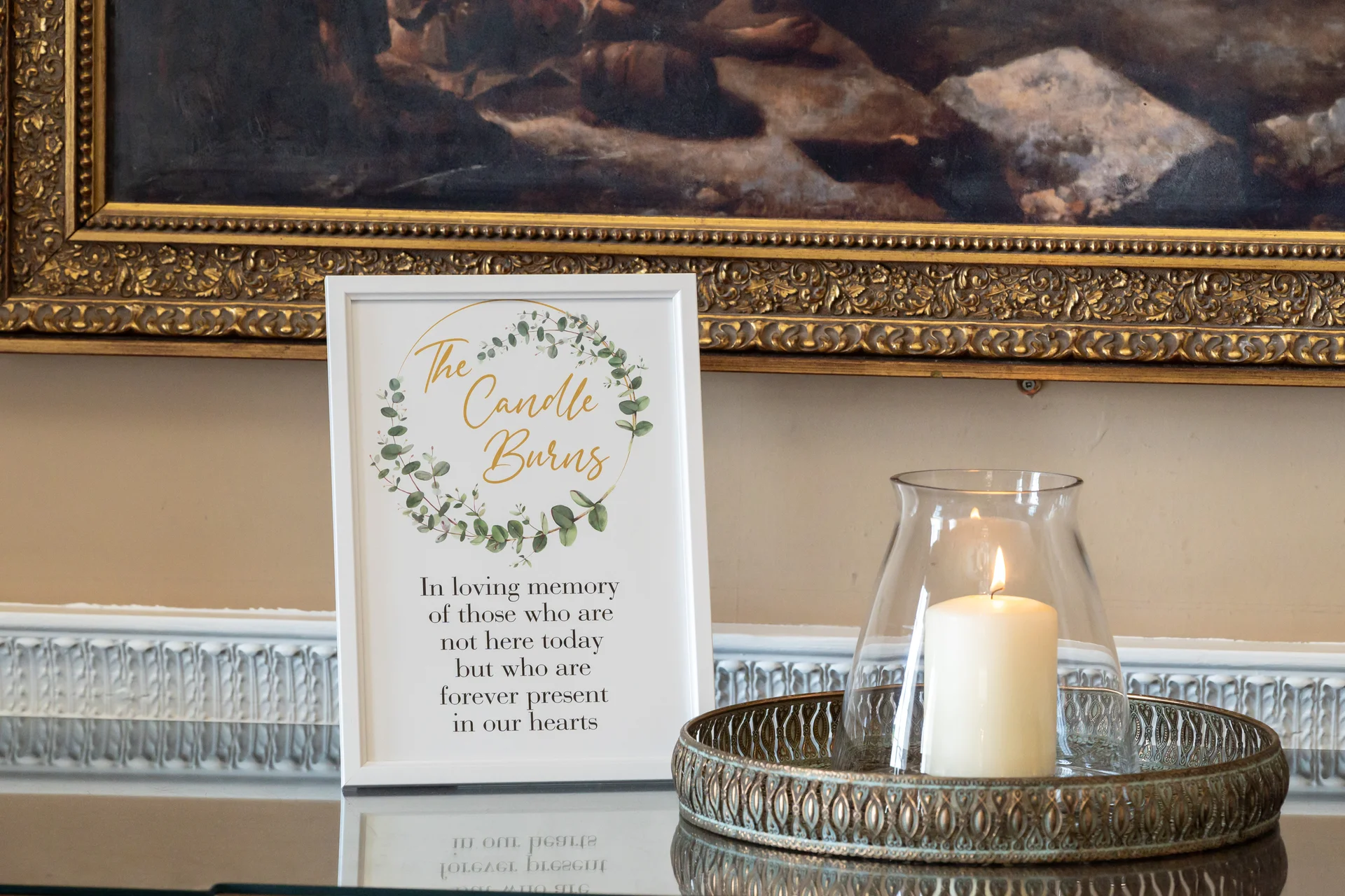 A framed memorial sign reading 'In loving memory' sits alongside a glowing white pillar candle inside a glass hurricane vase on a decorative gold tray.