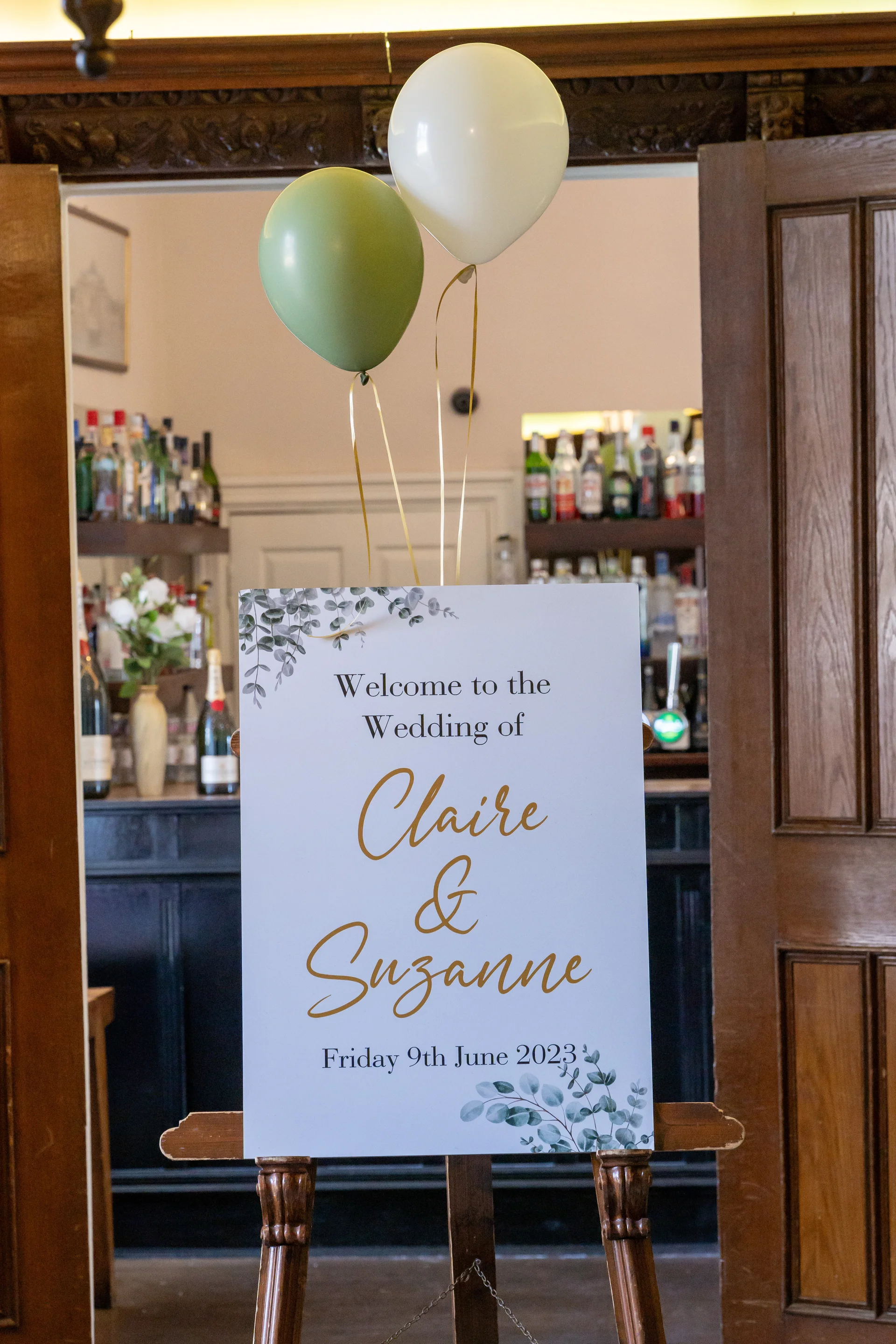An elegant wedding welcome sign positioned on a wooden easel features botanical eucalyptus details and gold lettering, topped with sage green and white balloons.