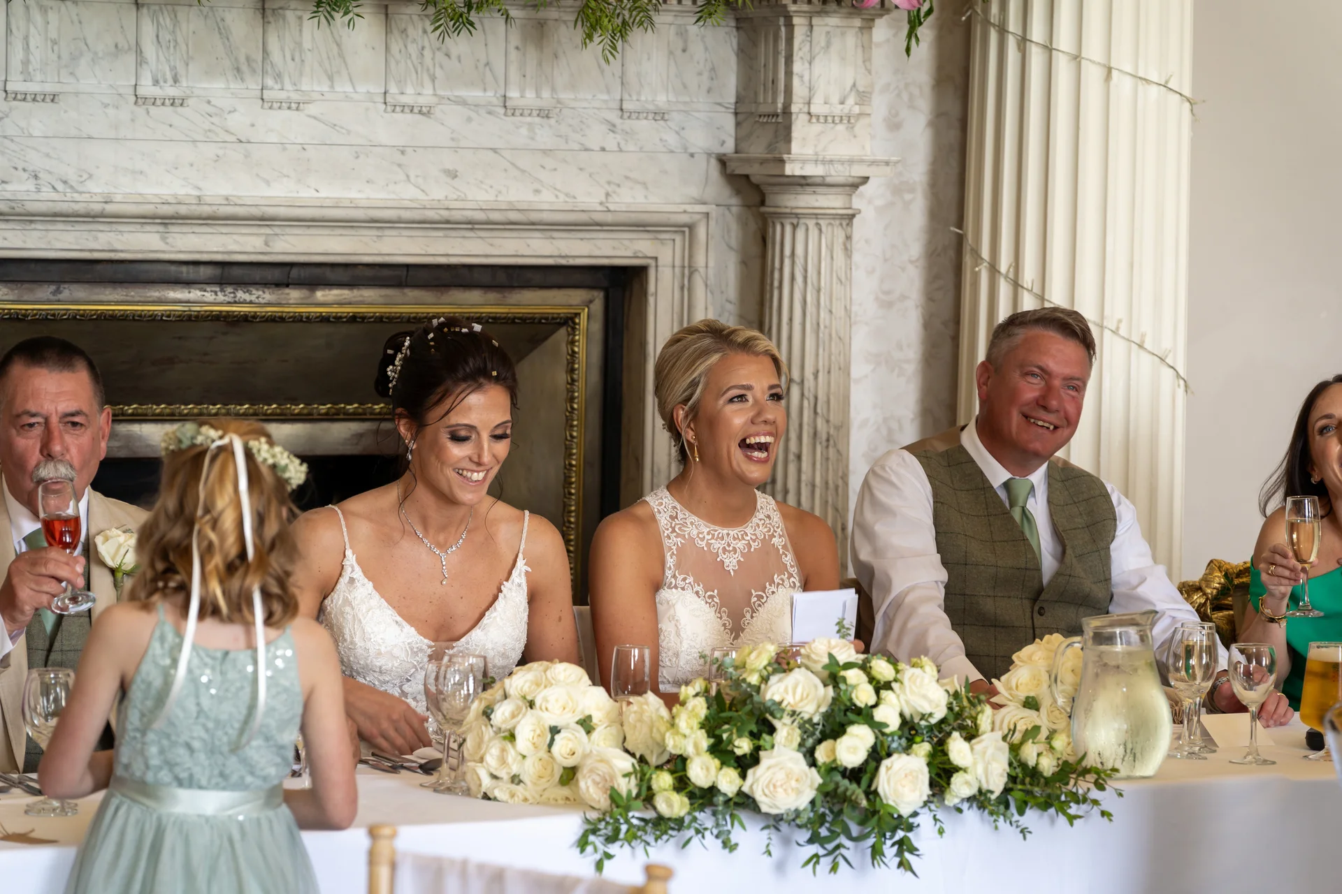A bridal party and guests laugh joyfully during the wedding speeches at a top table decorated with white roses in front of a marble fireplace.