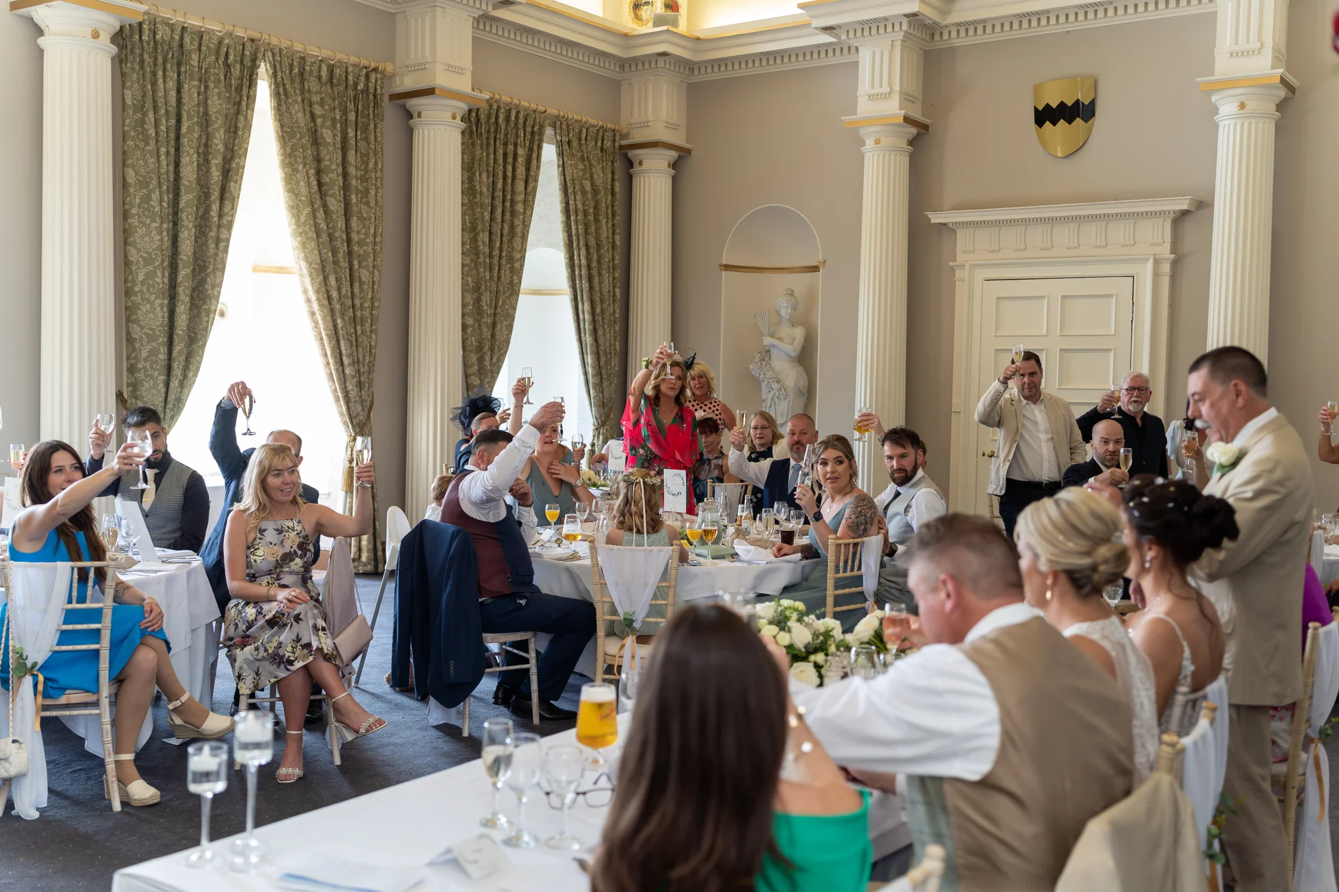 Wedding guests raise their glasses in a toast during the speeches inside a grand, classically decorated ballroom with tall pillars.